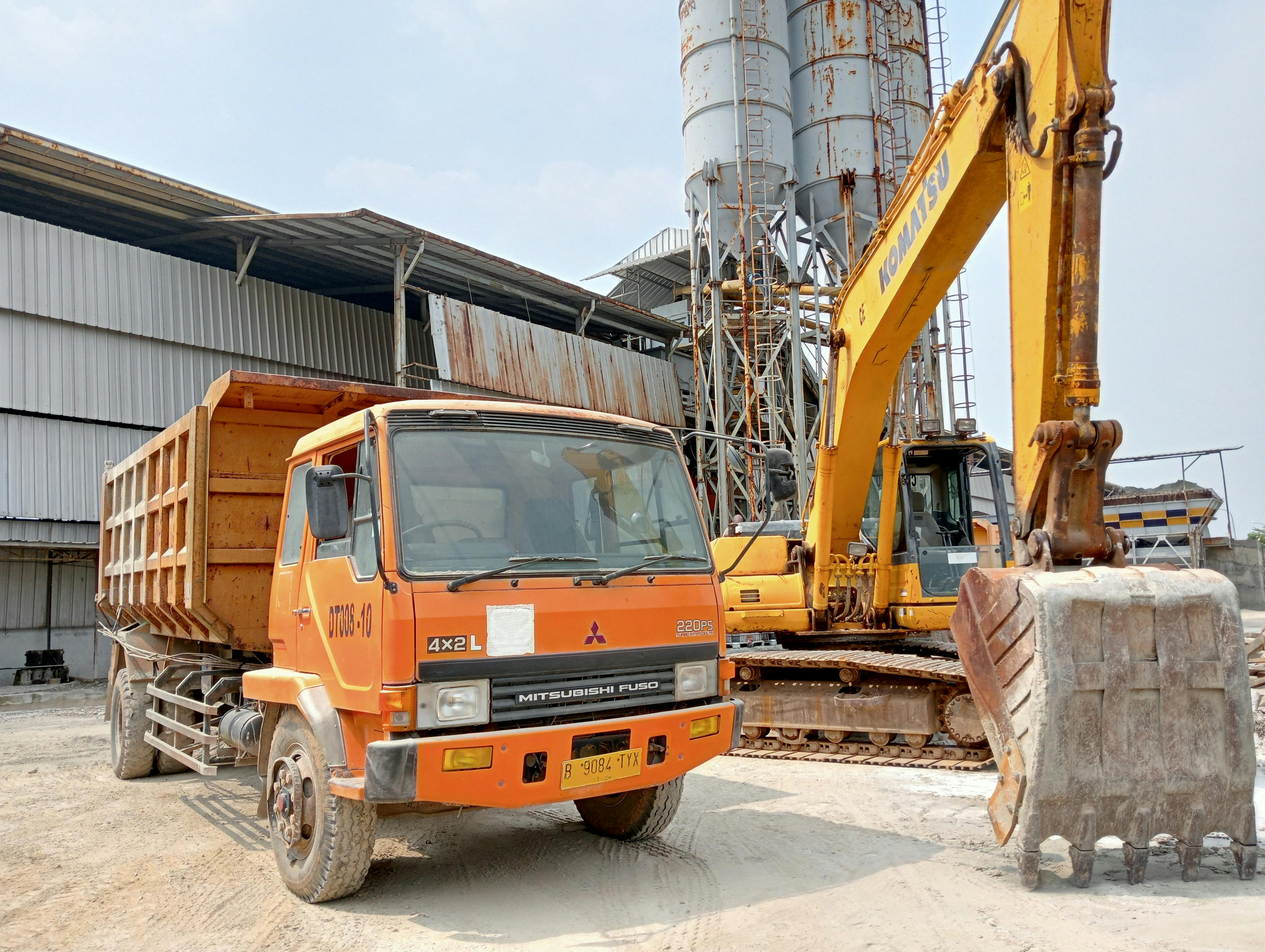 Construction site featuring a dump truck and excavator, with industrial buildings in the background.