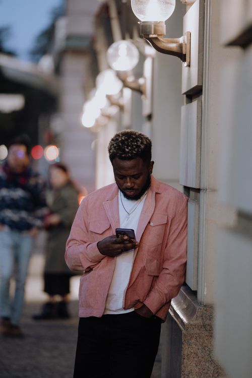 Free Stylish man leans against a building in Berlin at night, engrossed in his phone. Stock Photo