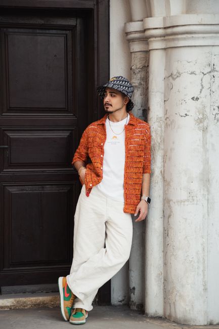 Man in orange shirt and wide pants leans on historic column, Adana.
