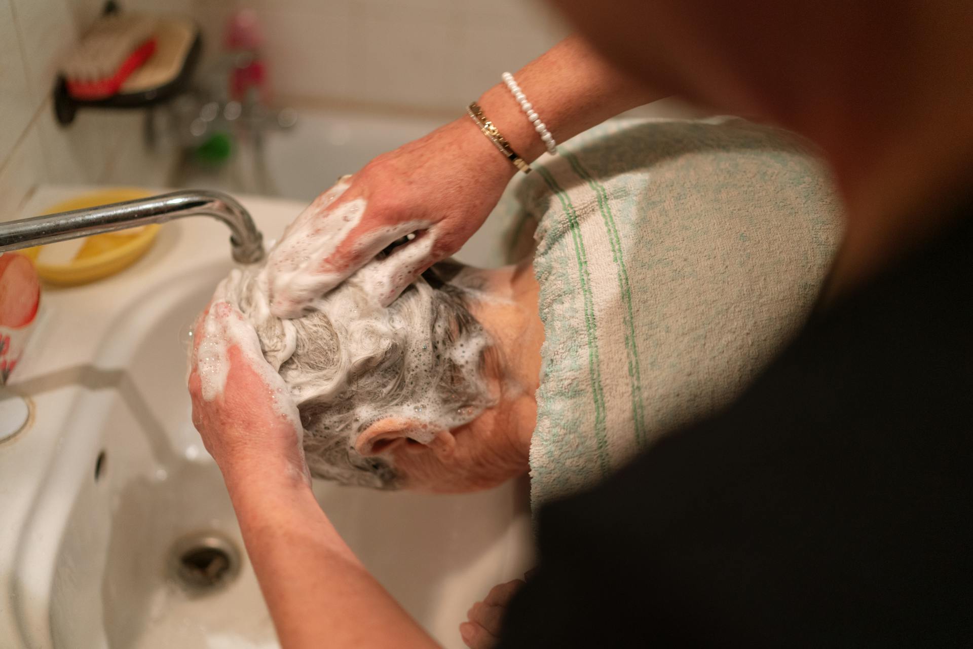 A person washing a senior woman's hair at a home sink, emphasizing care and grooming.