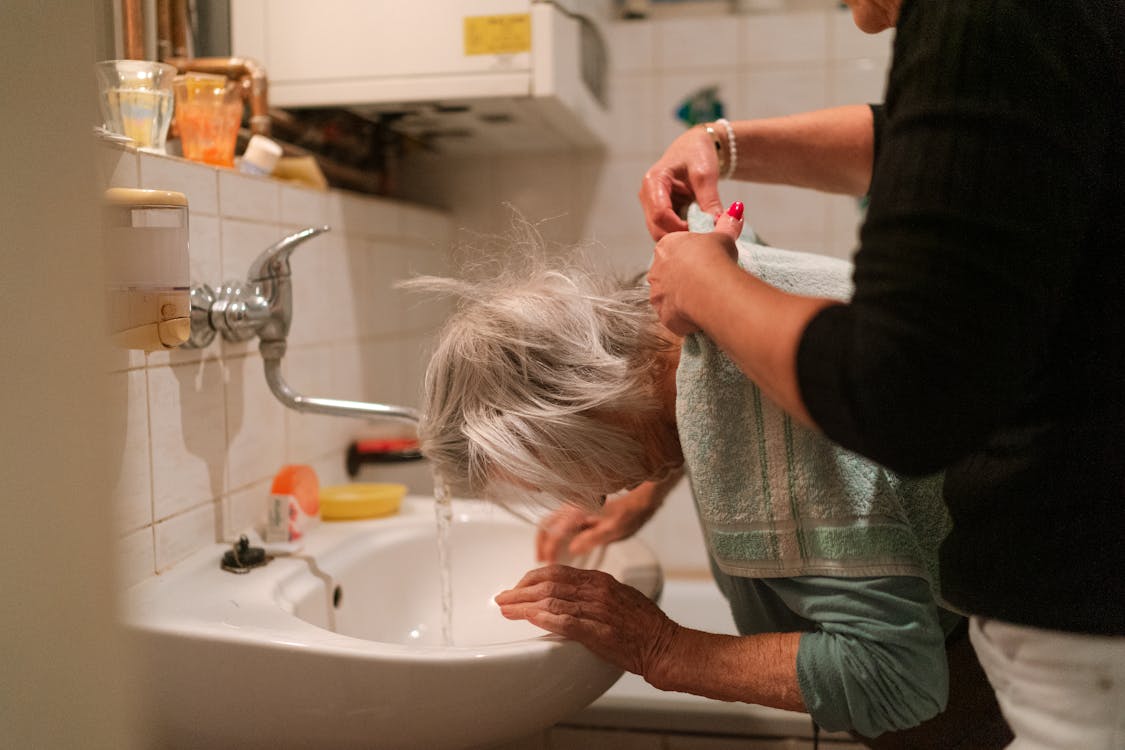 An old woman washing up in the sink with help from a caretakerin Dallas