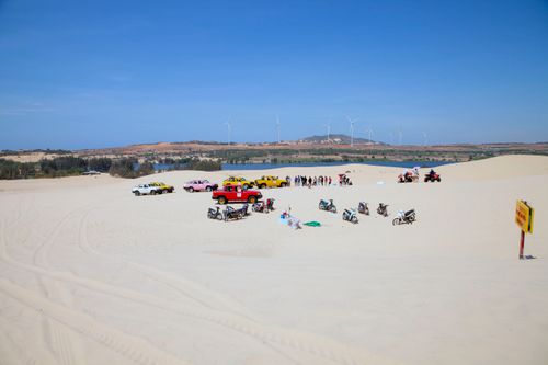 Free Tourists enjoy a sunny dune landscape with colorful vehicles and distant wind turbines. Stock Photo
