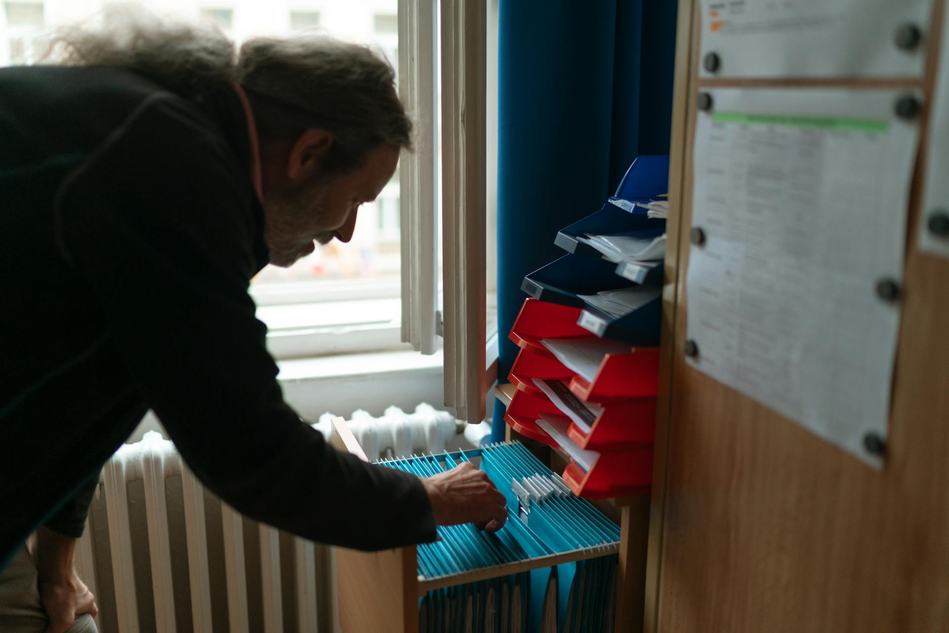 A man with long hair searches files in an organized office space.