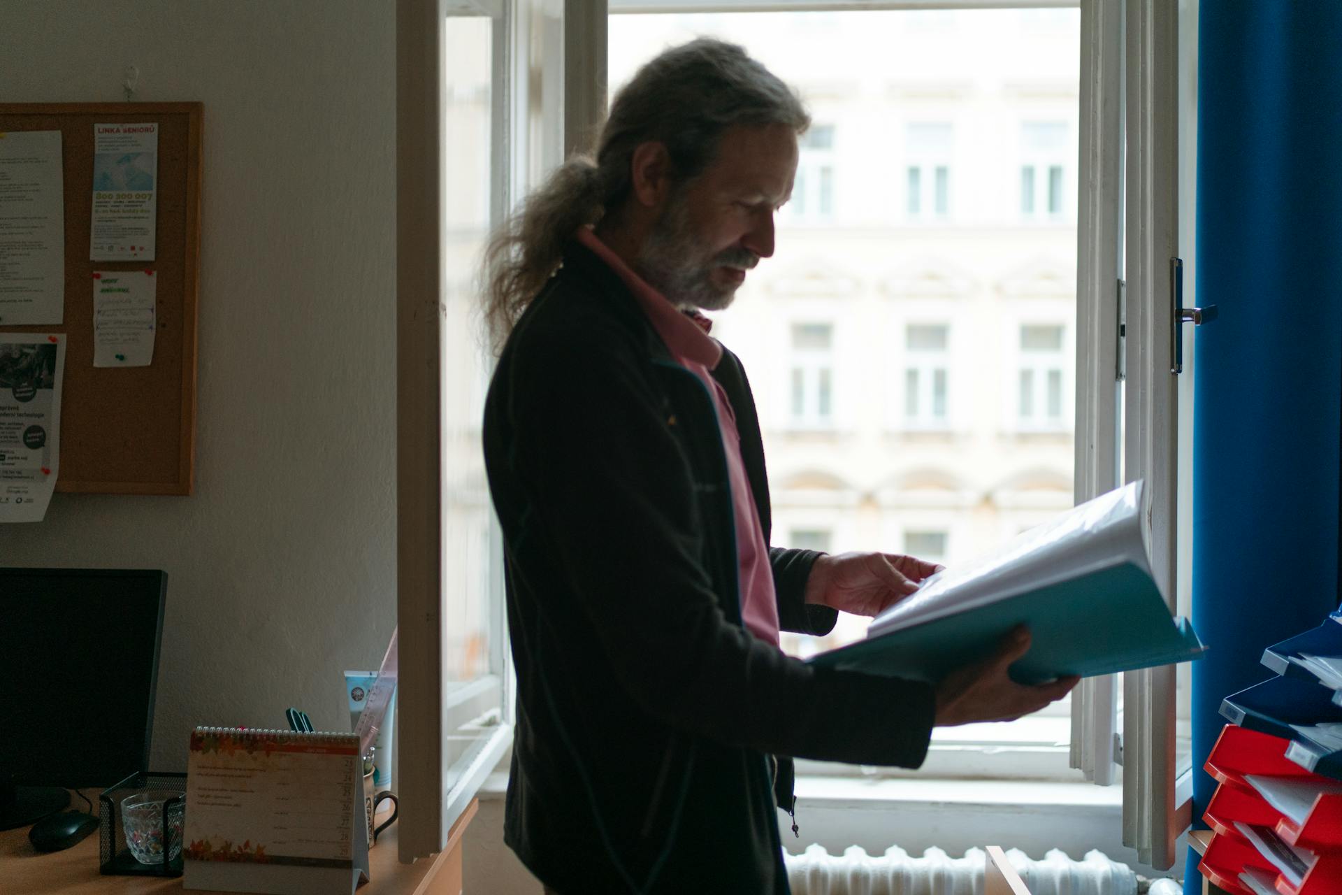 A senior man with long hair examines documents in a Prague office next to a window.