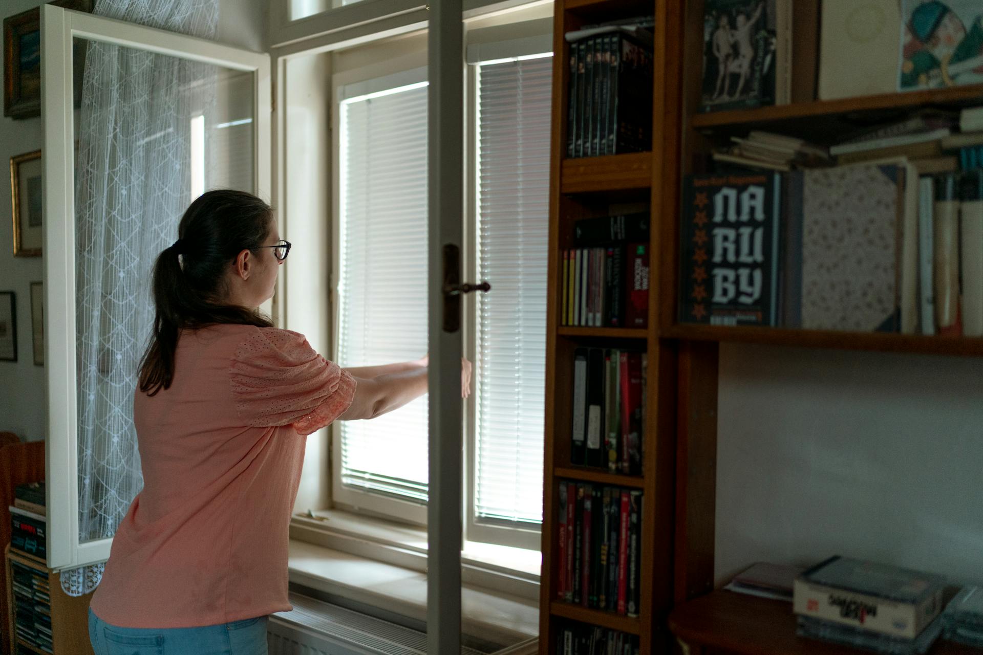 A woman in a casual pink blouse adjusts window blinds in a cozy home library.