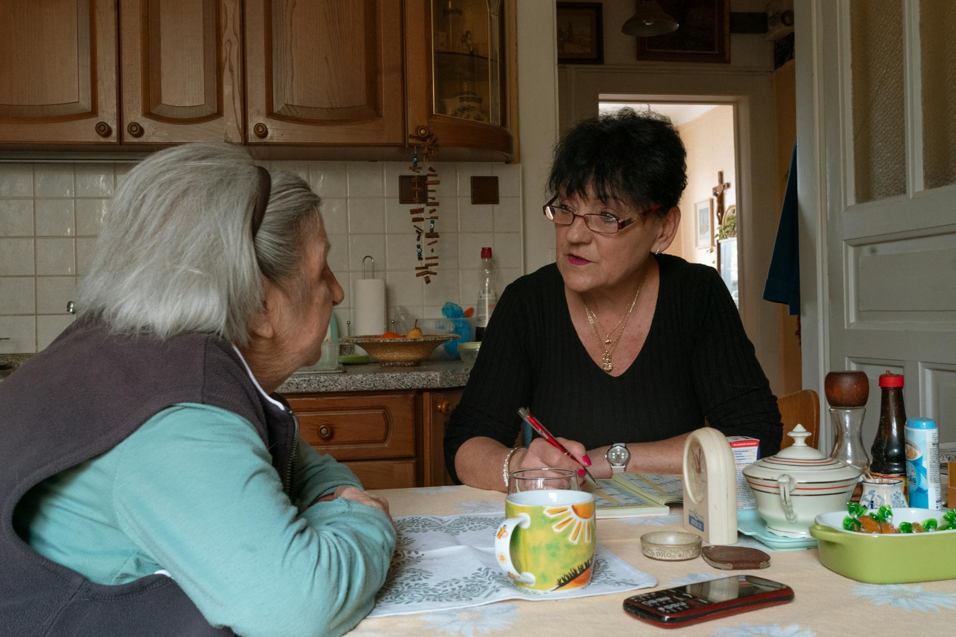 Senior woman and caregiver having a conversation in a cozy Prague kitchen setting.