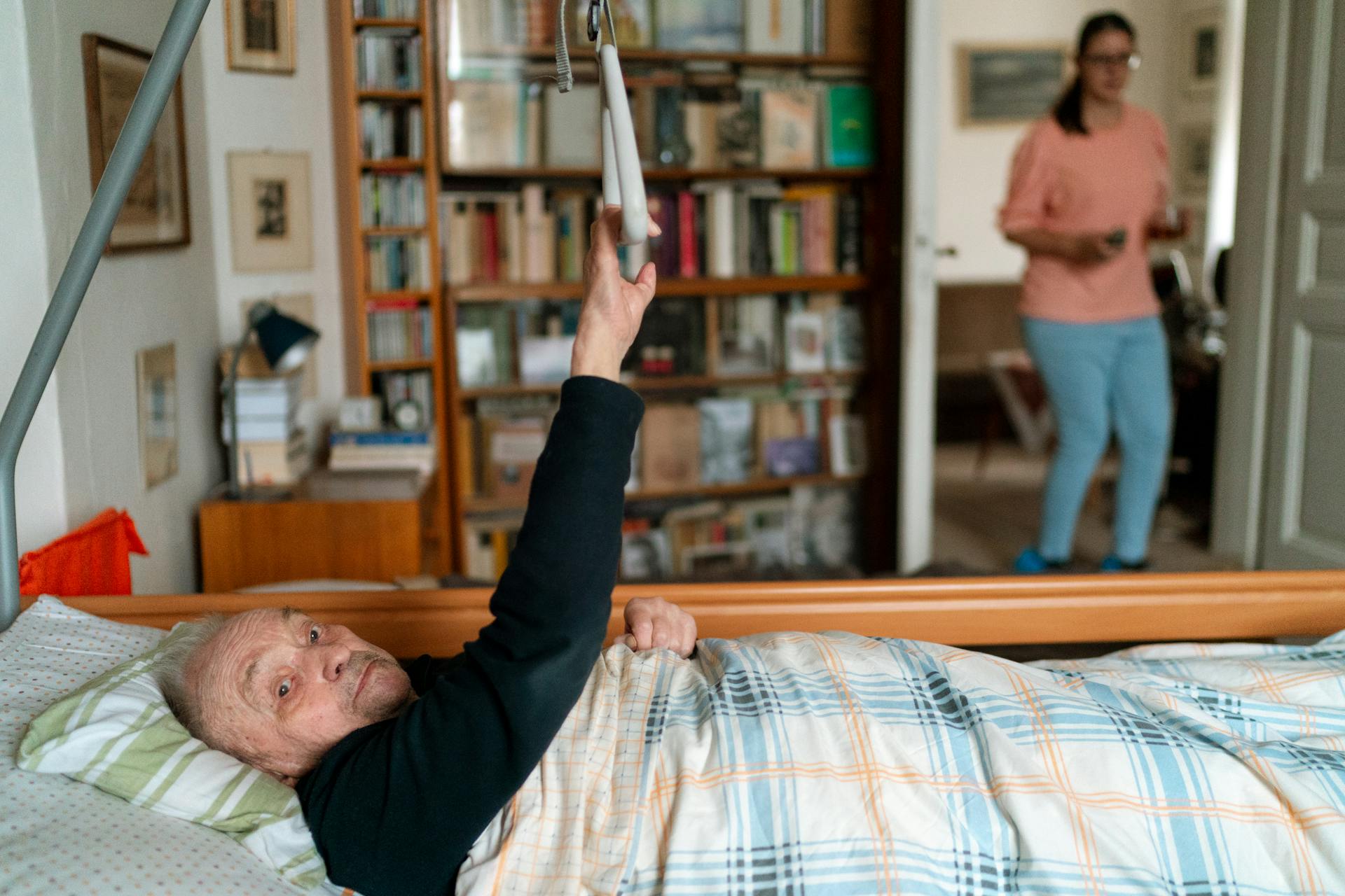 Senior man in bed with caregiver in Prague home, surrounded by books.