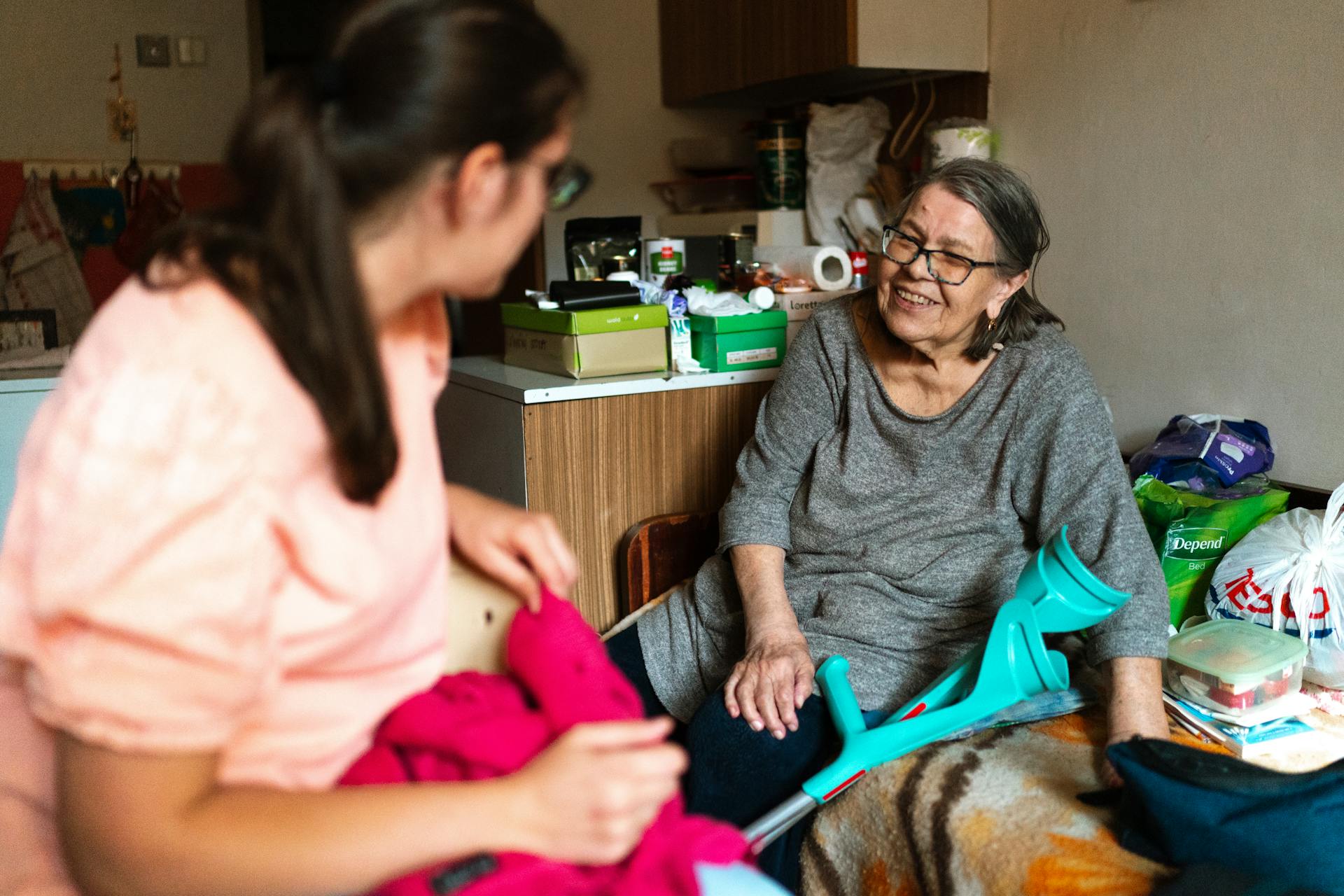 A caregiver helps a senior woman with a crutch in a cozy Prague home setting.