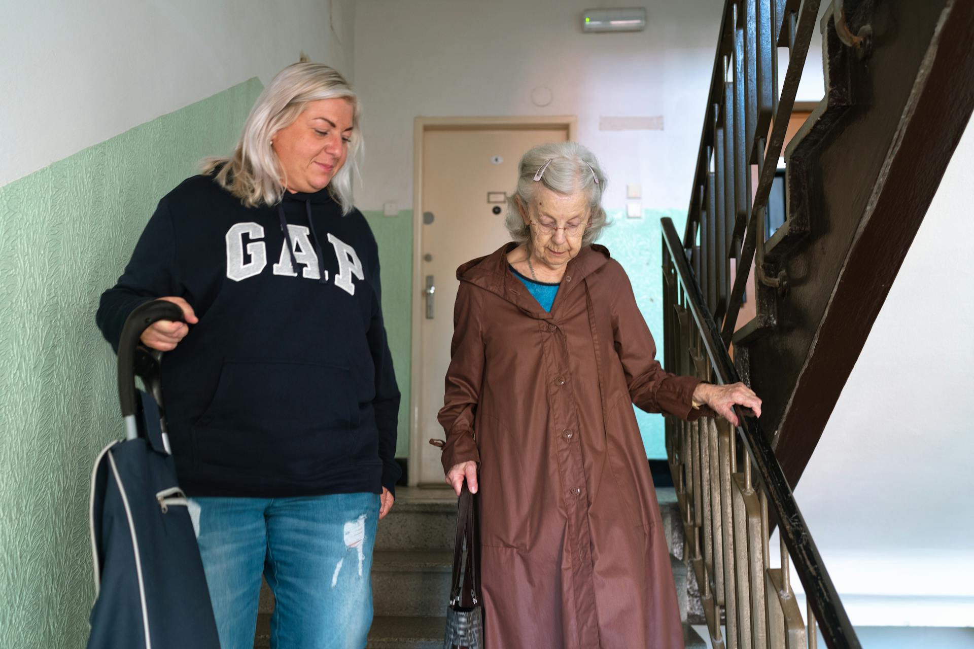 A senior woman assisted by a caregiver walking down indoor stairs in Prague.