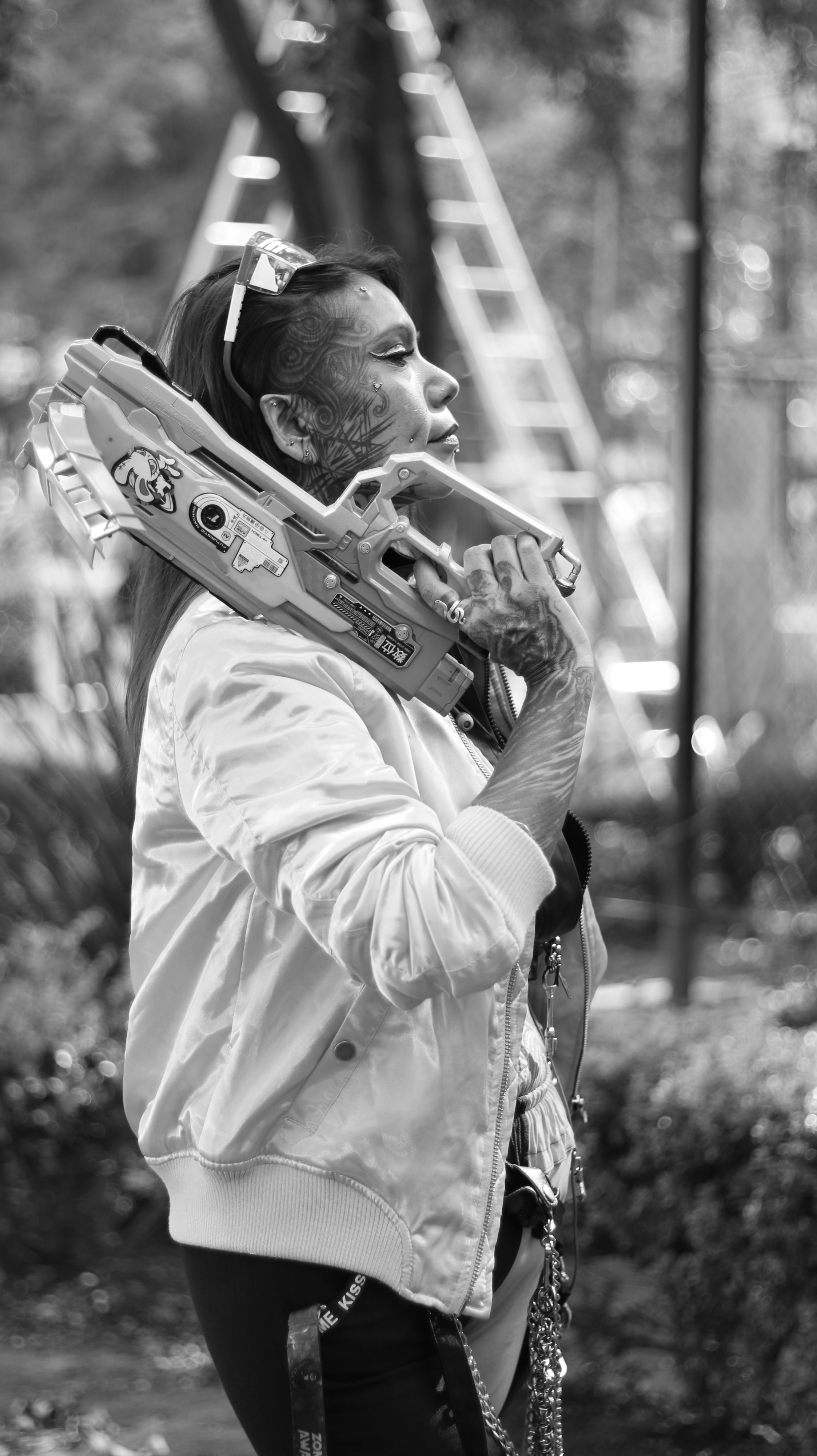Black and white portrait of a tattooed woman with a toy gun in an outdoor setting.