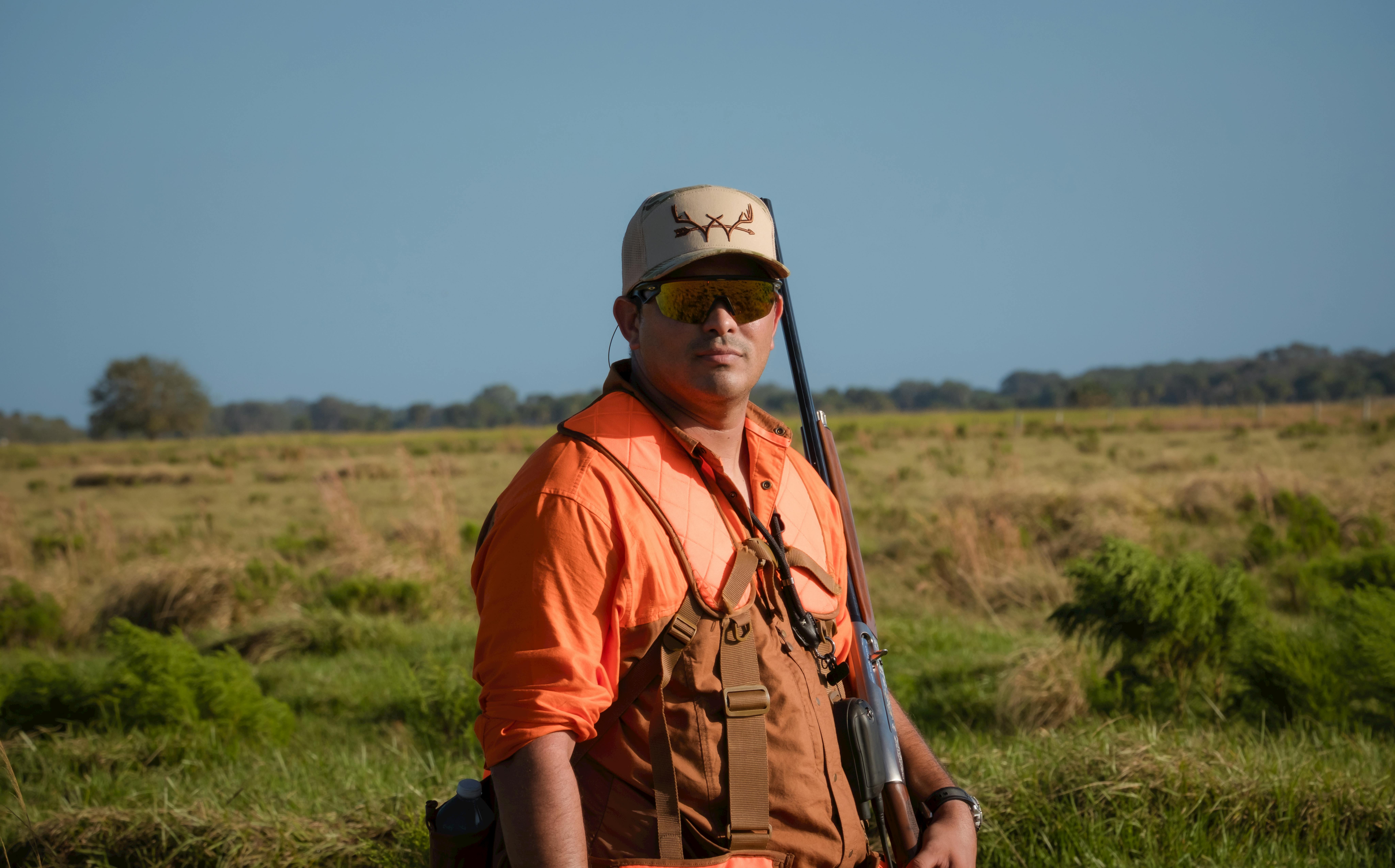 A hunter in orange gear poses in an open field under a clear sky
