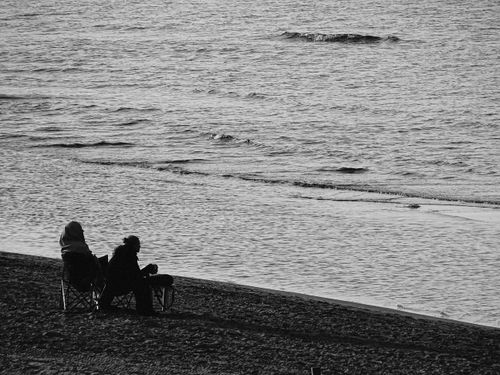Free Two individuals in chairs enjoying a peaceful moment by the sea in a black and white scene. Stock Photo