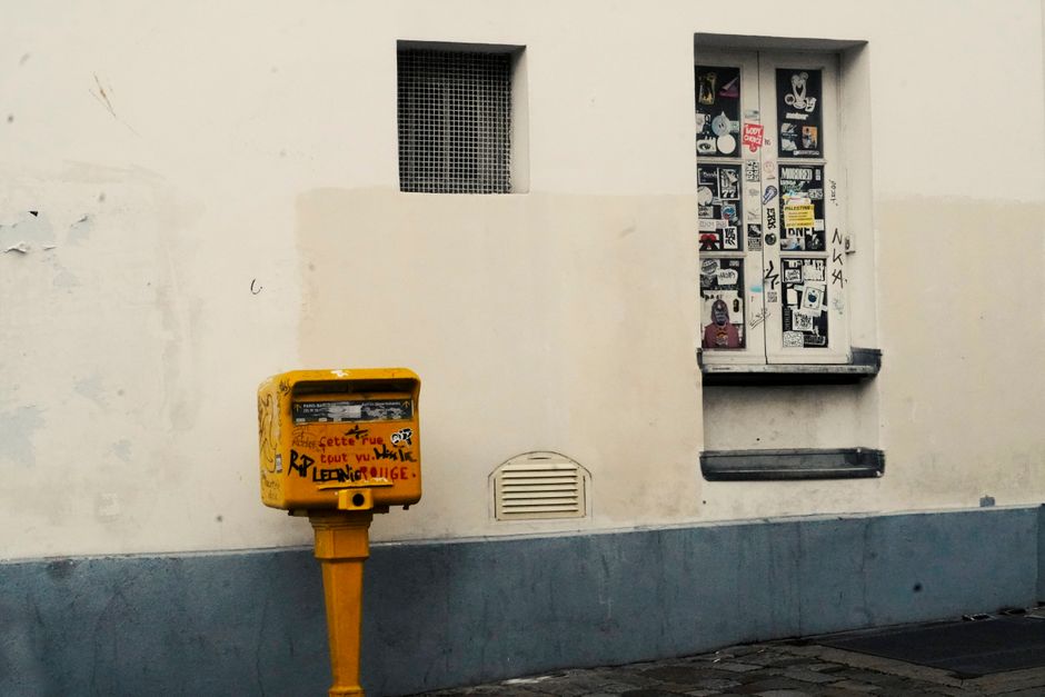 A yellow graffiti-covered mailbox on a street with sticker-plastered windows.