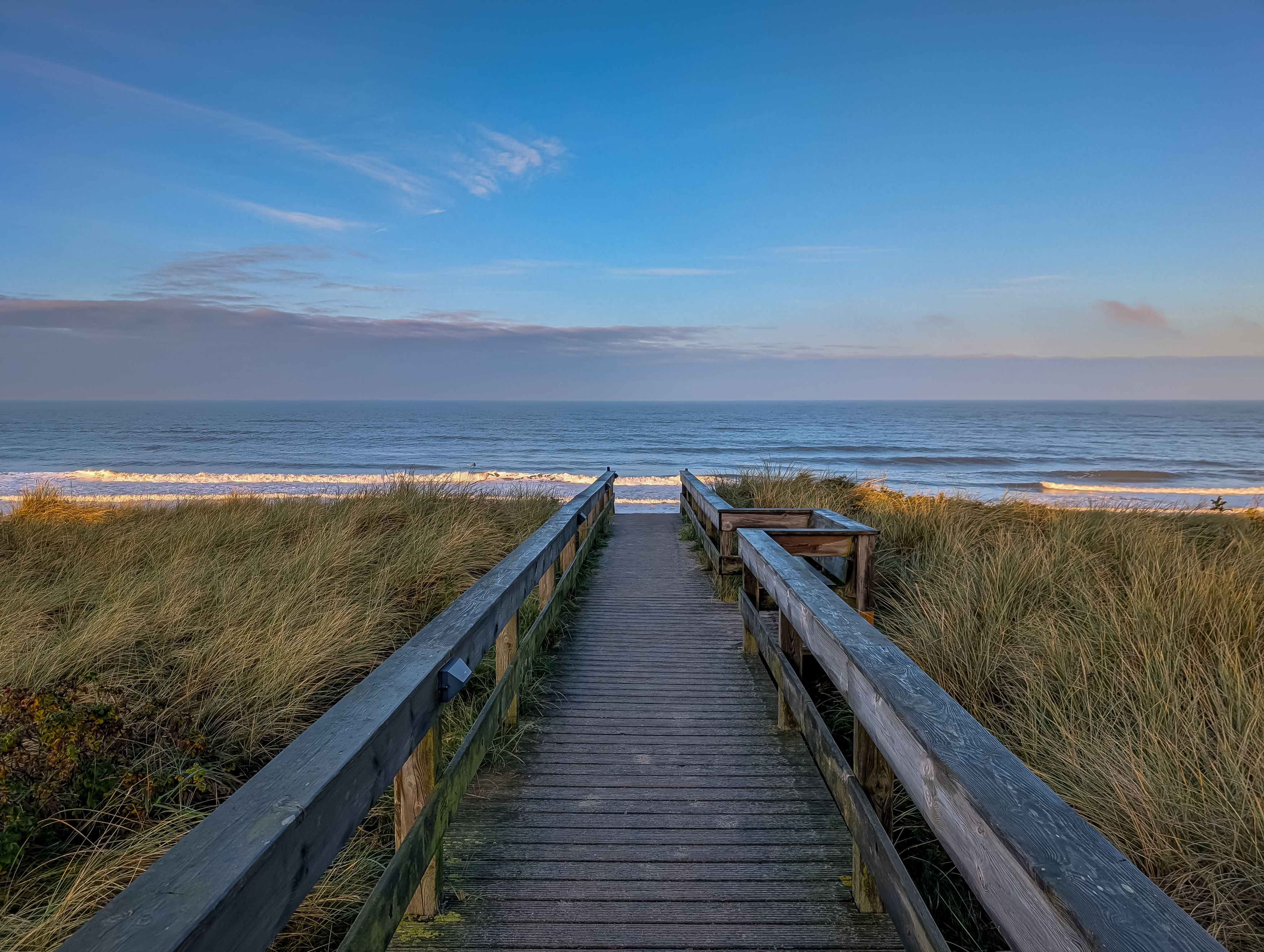 Wooden pathway leading to Sylt Beach with golden dunes and tranquil waves at sunset.