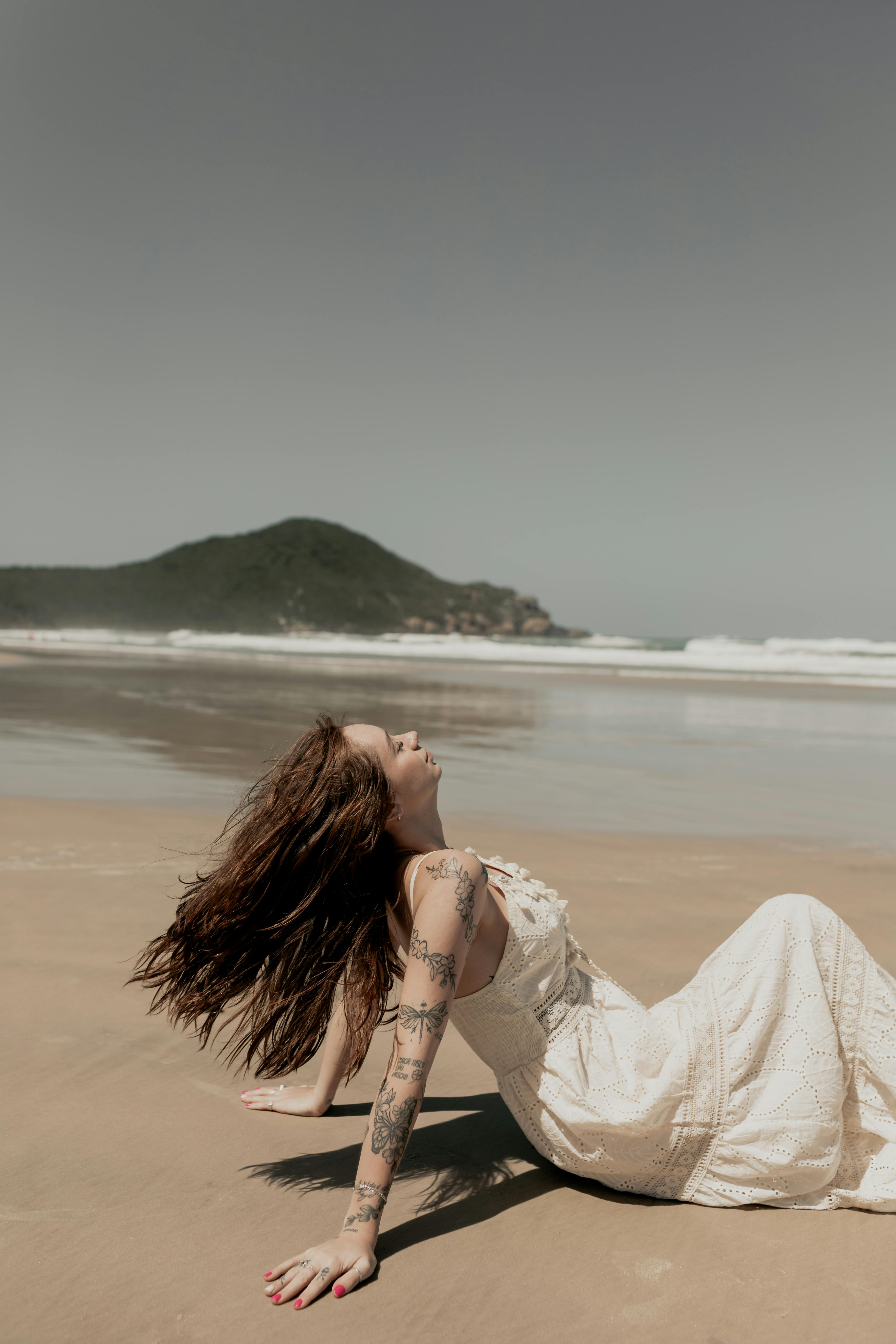 Woman in white dress enjoying the sun on a serene beach with waves and mountains.