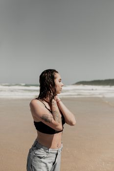 A woman with tattoos stands thoughtfully on a sunny beach, enjoying the ocean breeze.
