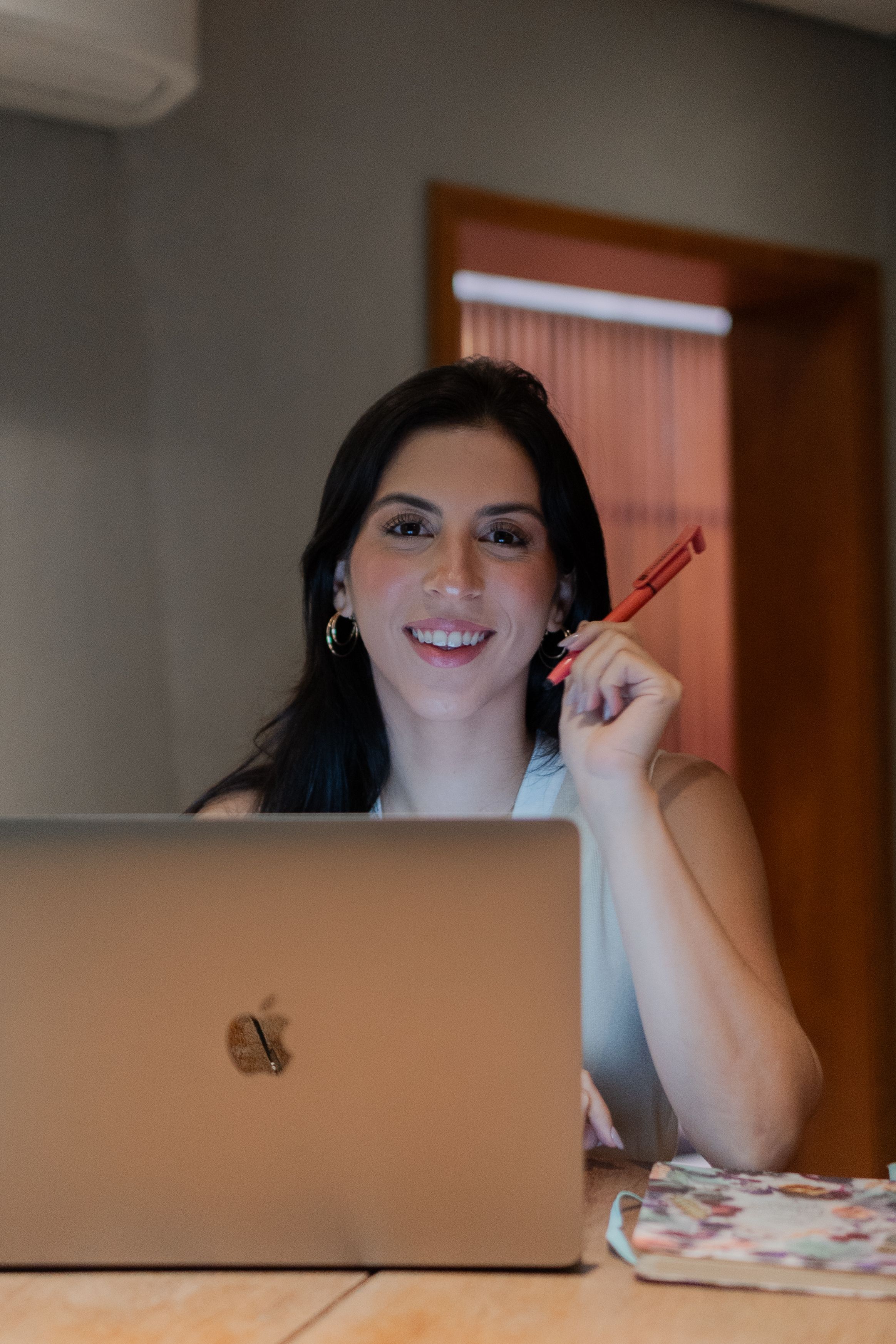 Smiling woman holding pen while working on laptop at desk.