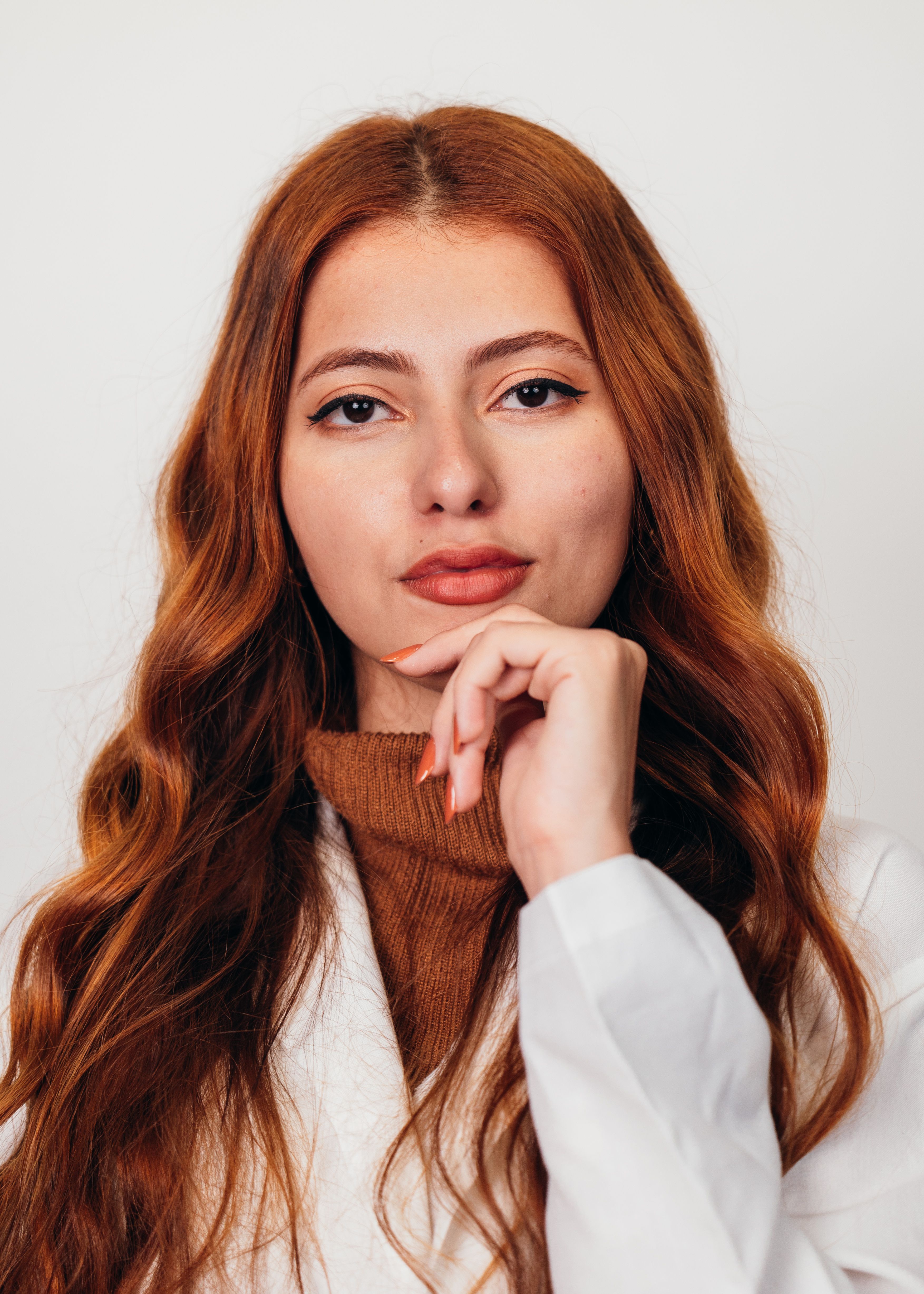 A close-up portrait of a confident young woman with long red hair wearing a white coat and brown sweater.