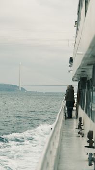 Person standing on ferry deck with bridge in distance over calm sea.