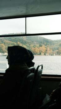 Silhouette of a woman gazing out of a ferry window at a scenic river landscape.