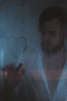 A man stands behind a fogged-up shower door, drawing a heart on the glass.