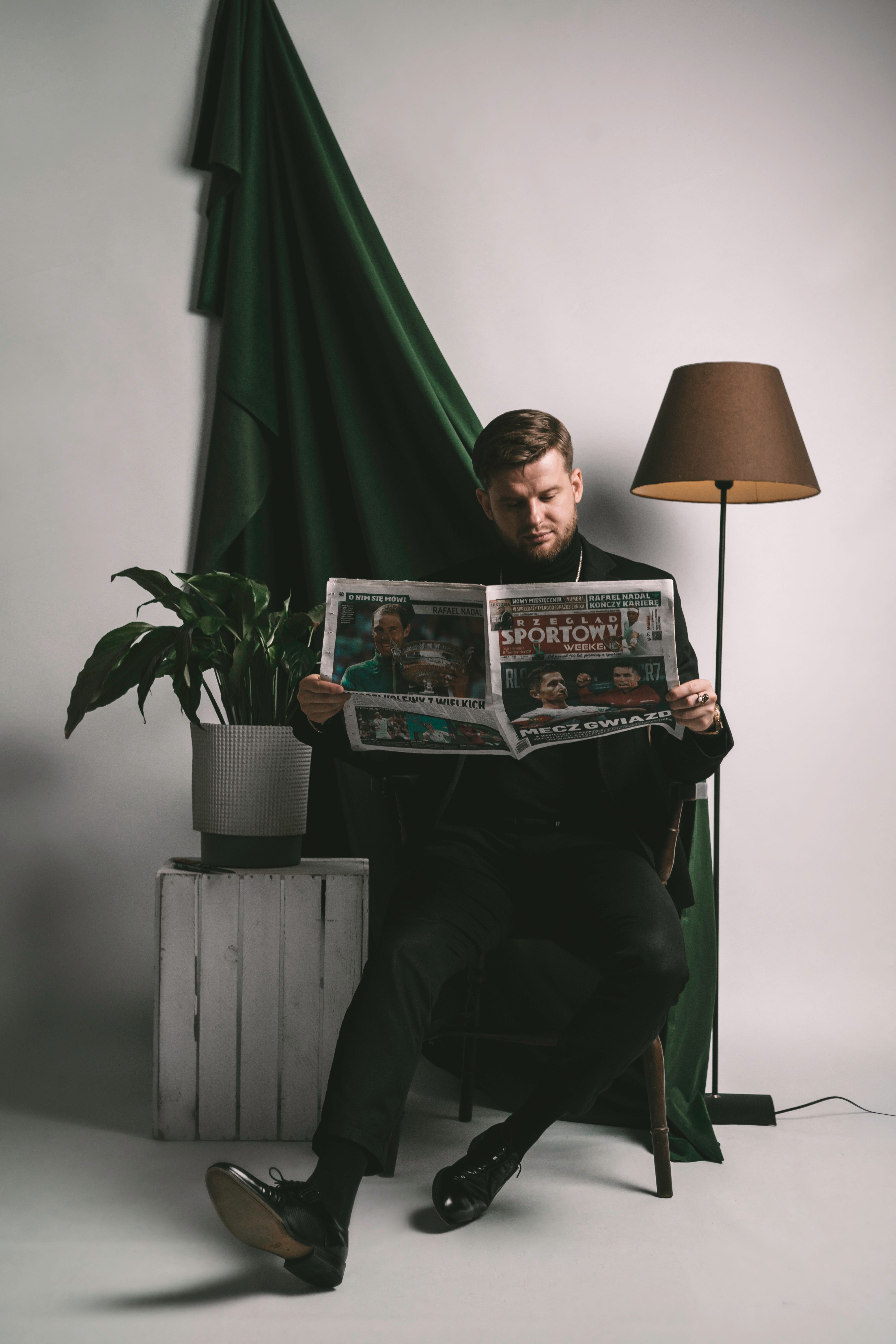 A well-dressed man sits elegantly on a chair reading a newspaper in a sophisticated, minimalist setting.