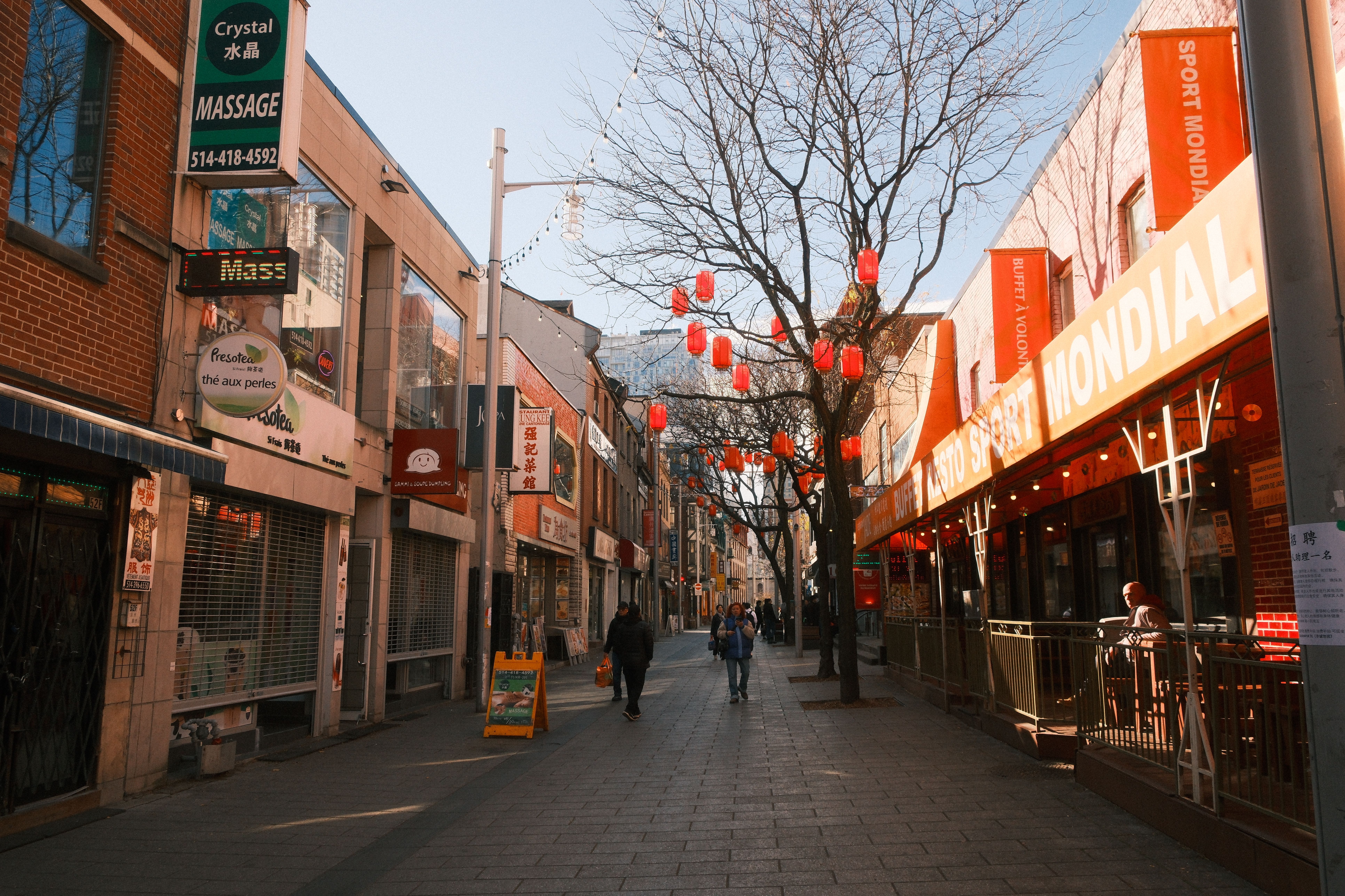 Chinatown street scene with red lanterns under trees, shops line both sides on a sunny day.