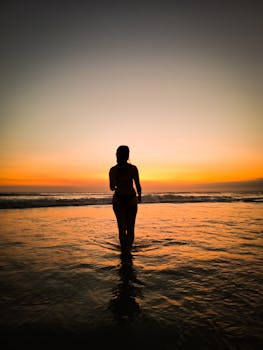 A woman stands in silhouette against a vibrant beach sunset, creating a tranquil scene.