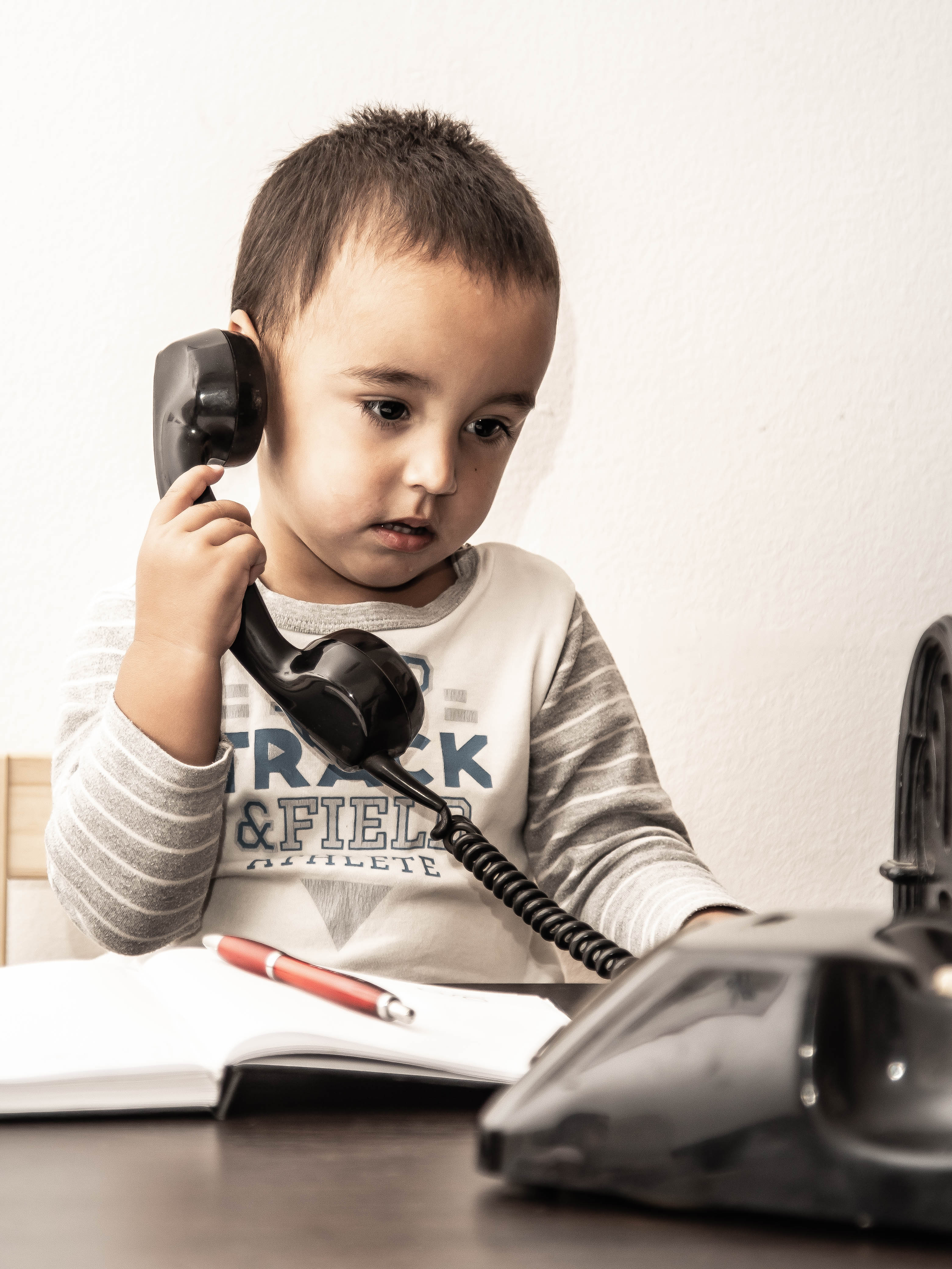 A young child using a vintage rotary phone while writing indoors.