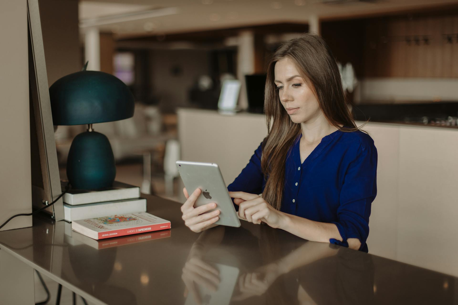 Woman reading on a tablet at a desk with books in a modern office setting.