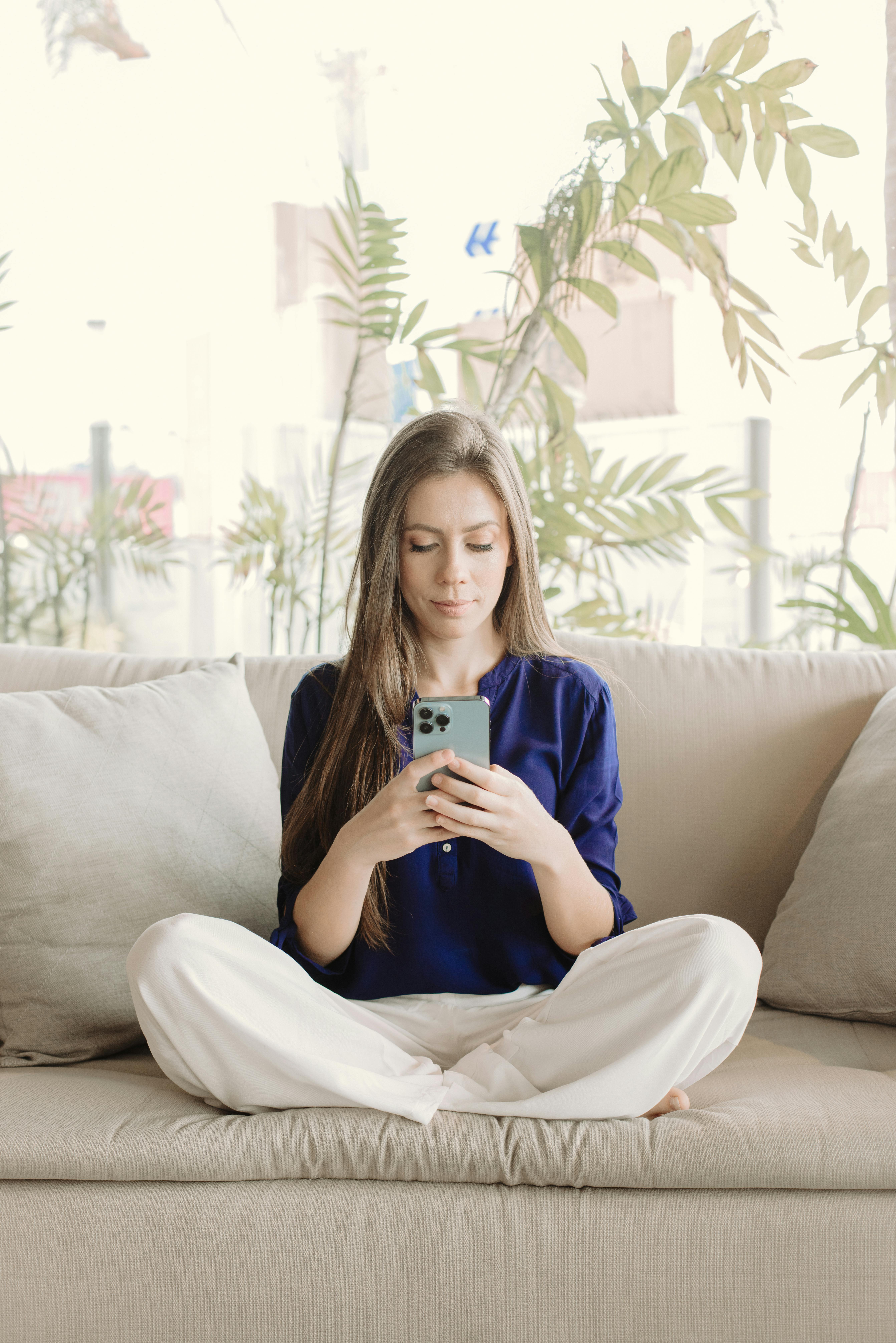 A young woman in casual attire sits on a couch using her smartphone in a bright indoor setting.