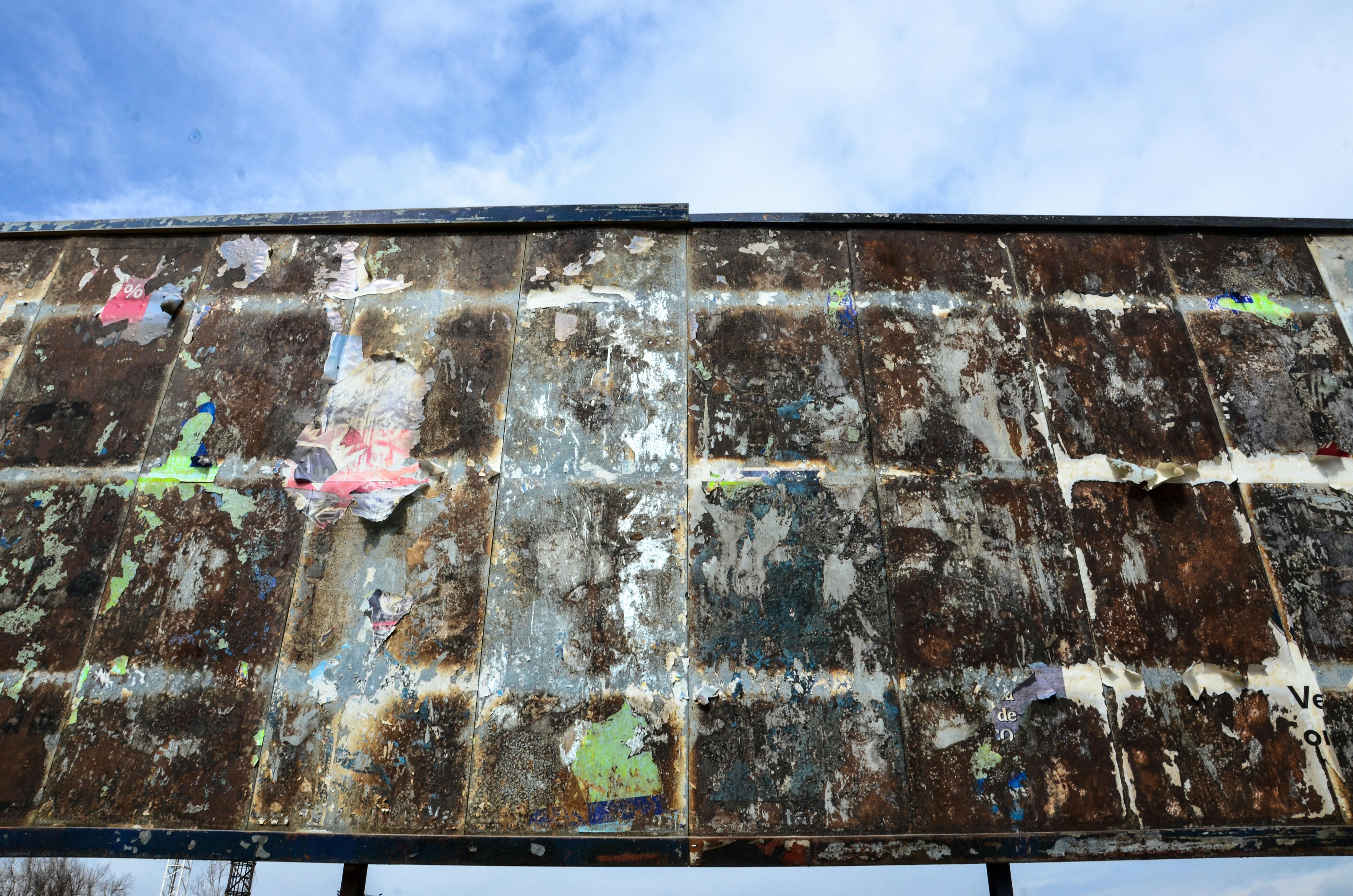 A rusty and torn billboard panel against a clear blue sky, evoking urban decay.