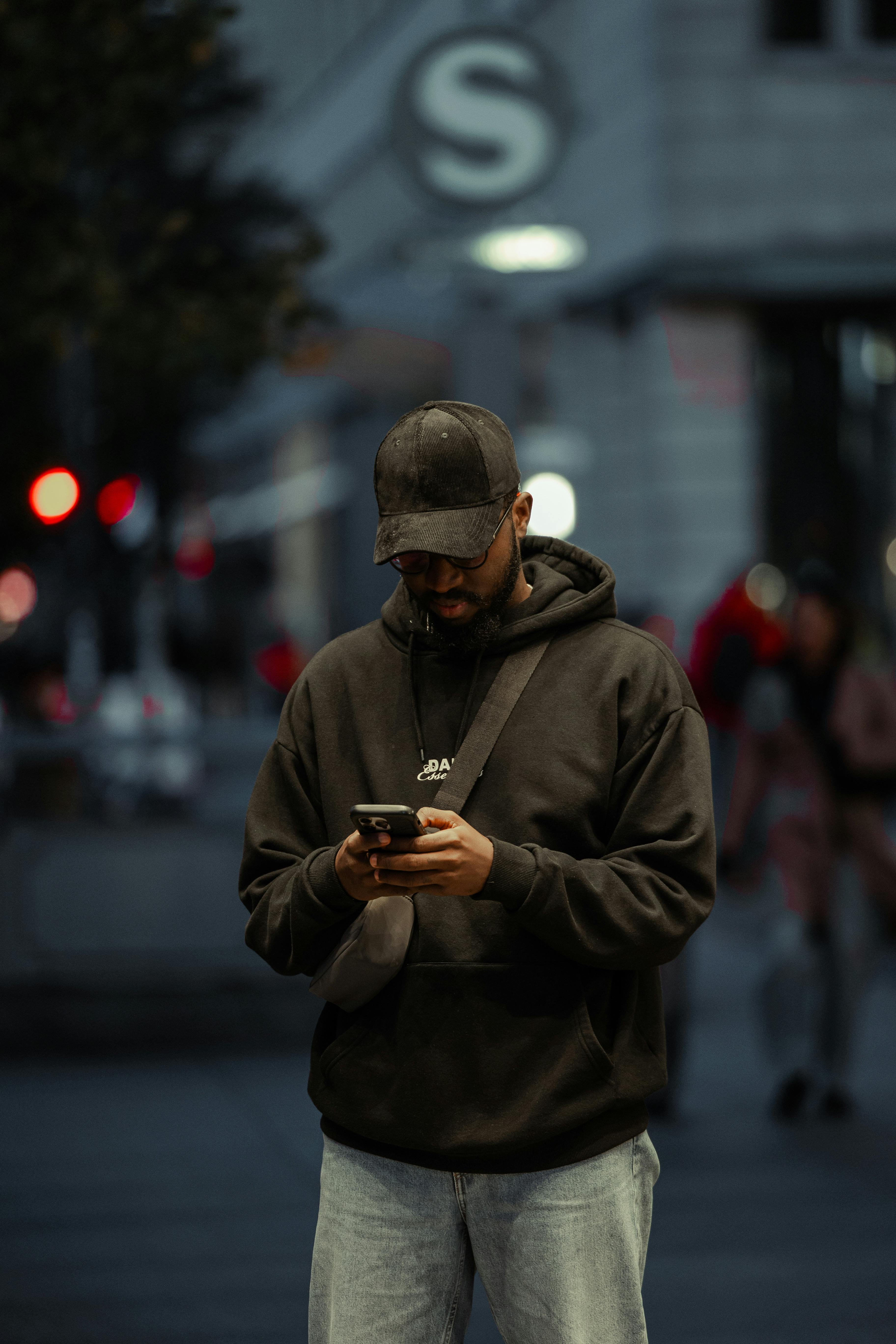 A man in a black hoodie checks his phone on a city street at night, surrounded by city lights.