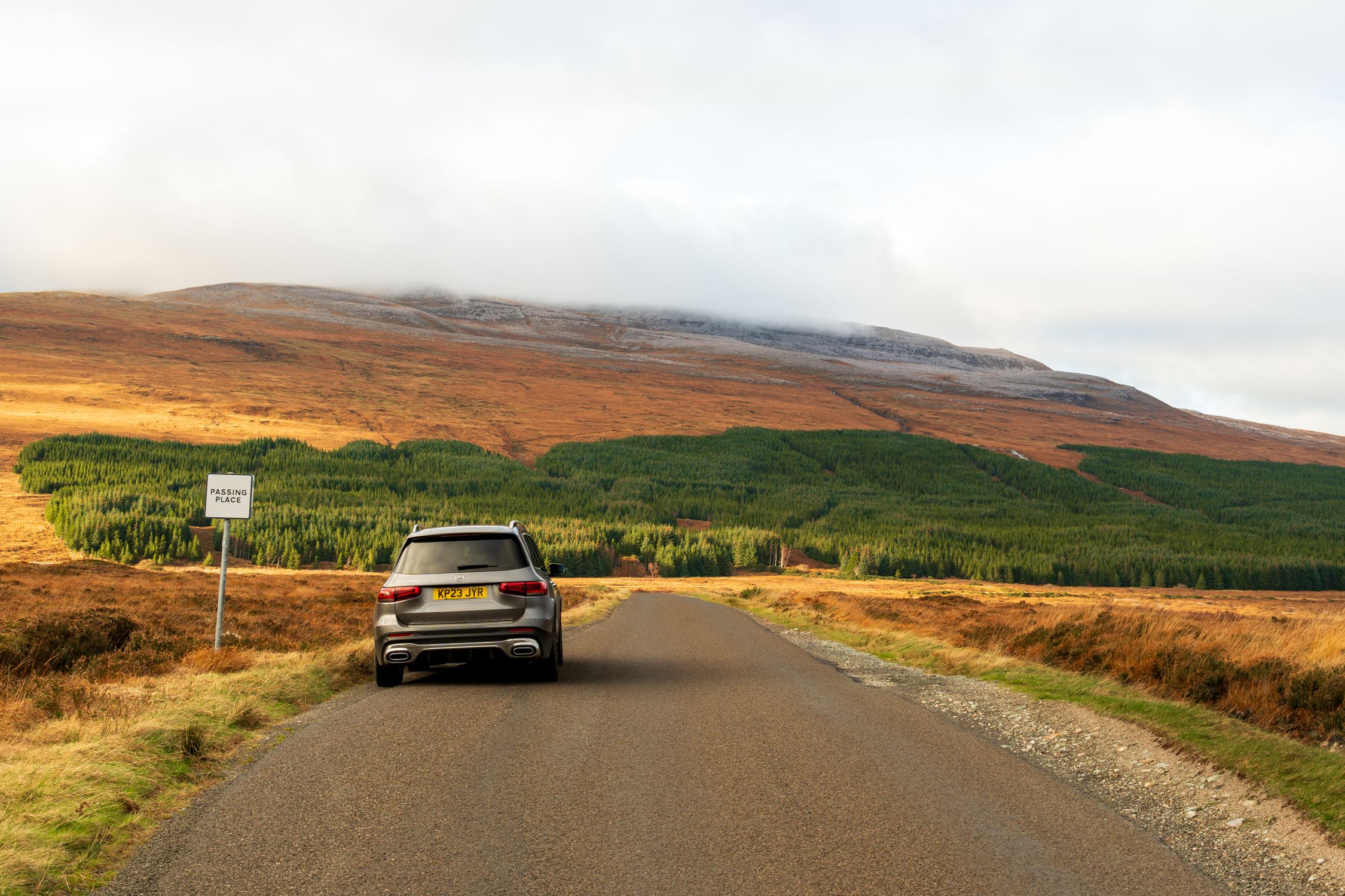 Car on a scenic road in the Scottish Highlands with lush green forests and rolling hills.