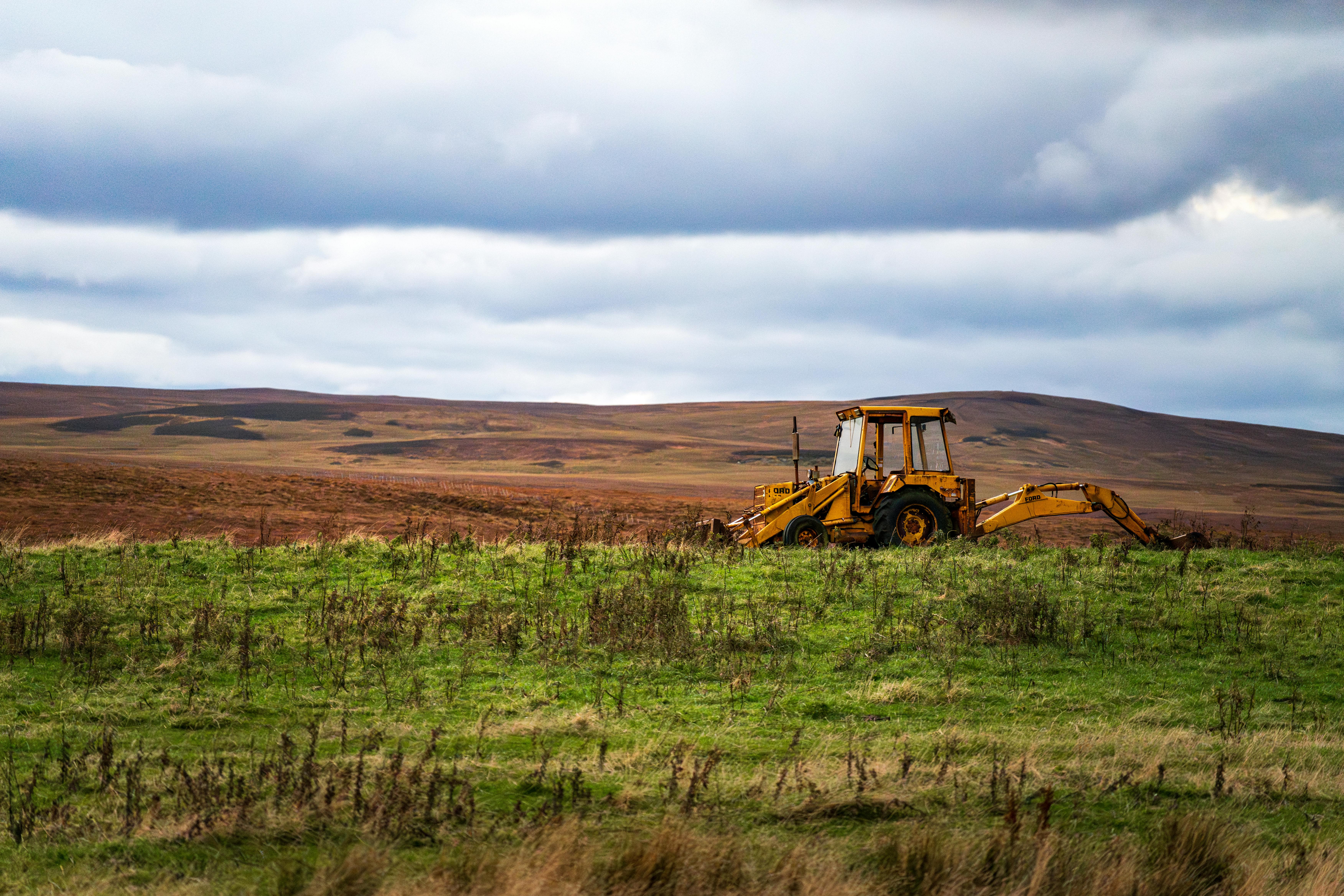 yellow backhoe on vast green field under cloudy sky