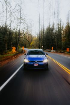 A blue car speeds down a scenic forest road, showcasing motion and autumn scenery.