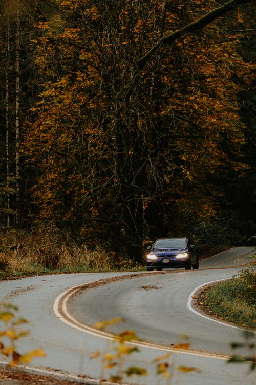Free A car navigates a curved road through a dense forest with vibrant autumn leaves. Stock Photo
