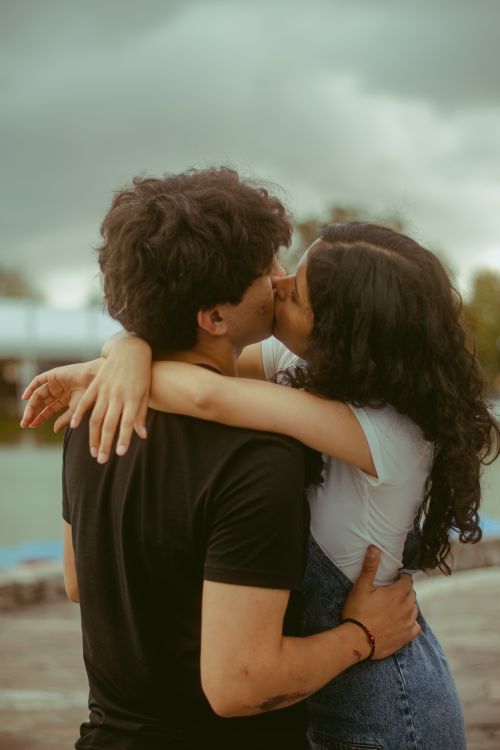 Free A romantic moment of a young couple kissing by the water on a cloudy day. Stock Photo
