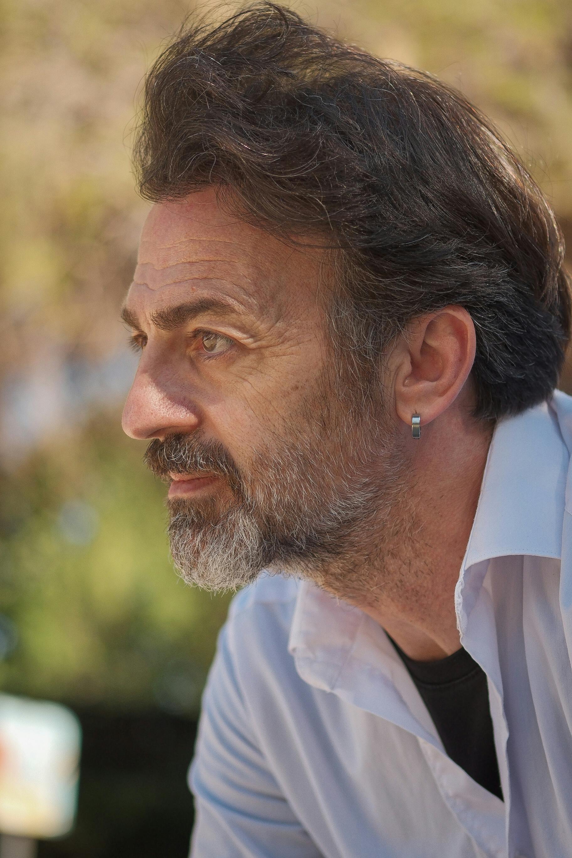 A contemplative senior man with a beard outdoors in sunlight.