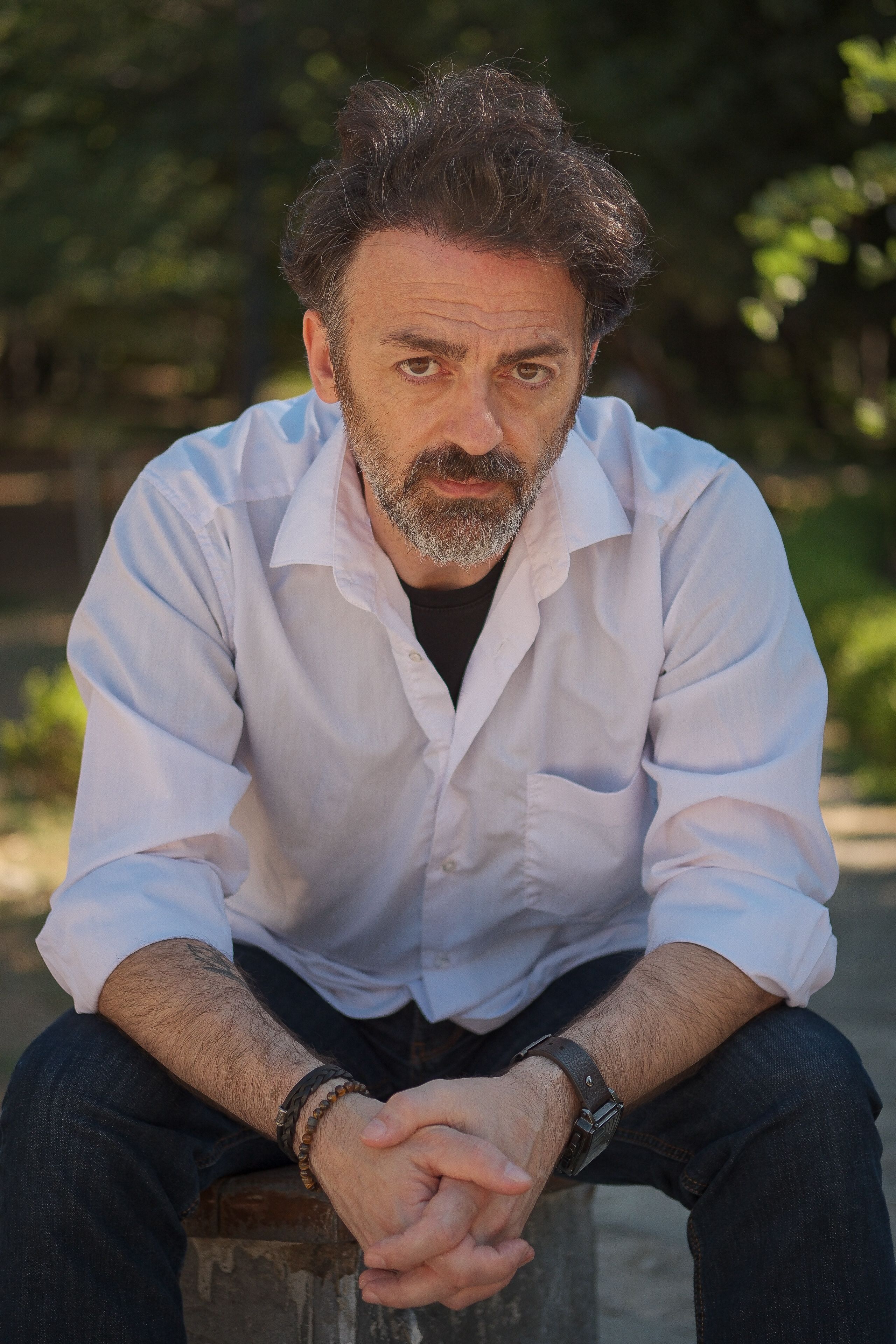 A thoughtful man sitting outdoors in a casual white shirt with greenery in the background.