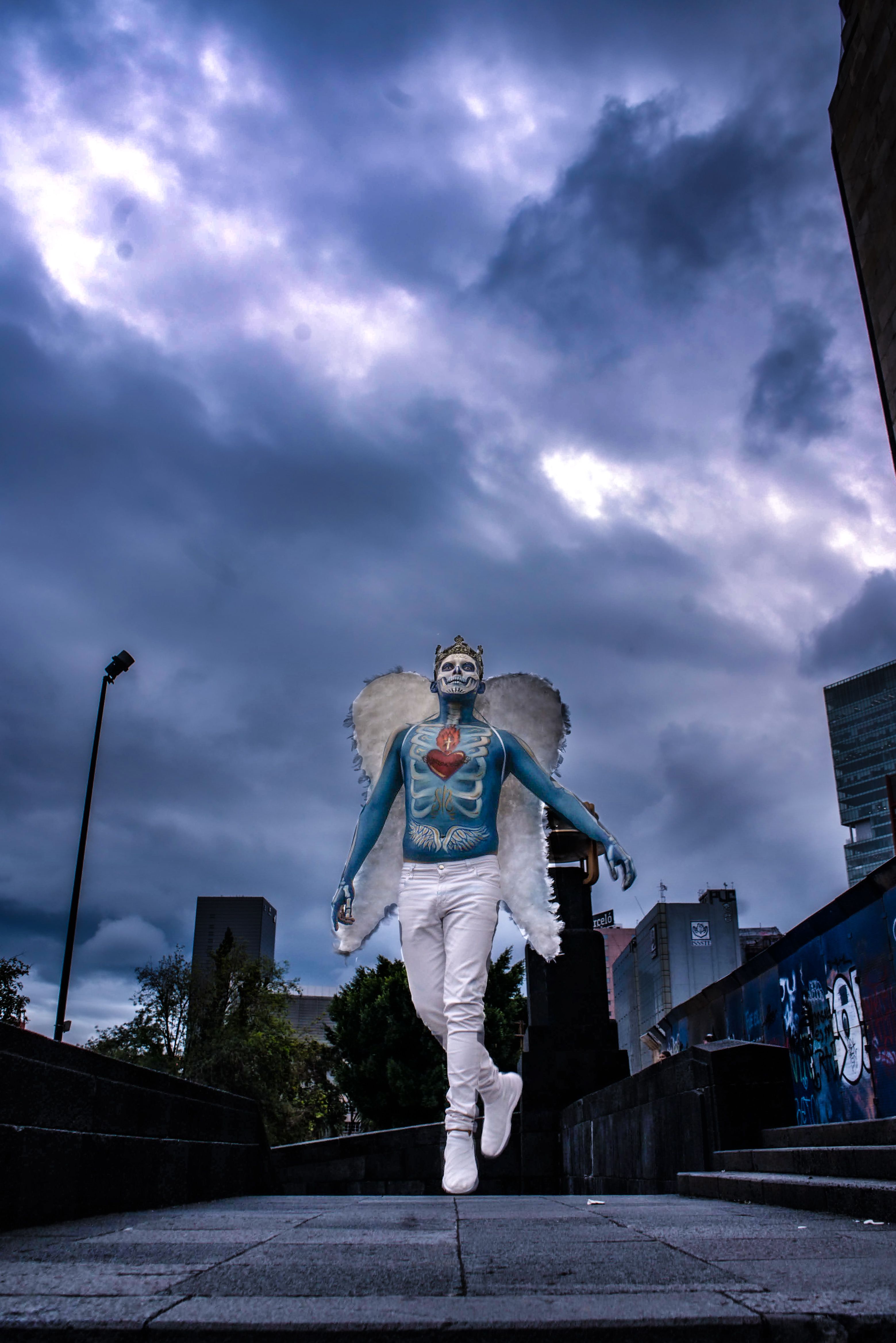A person in Catrina makeup and angel wings walks under a stormy sky in Mexico City.
