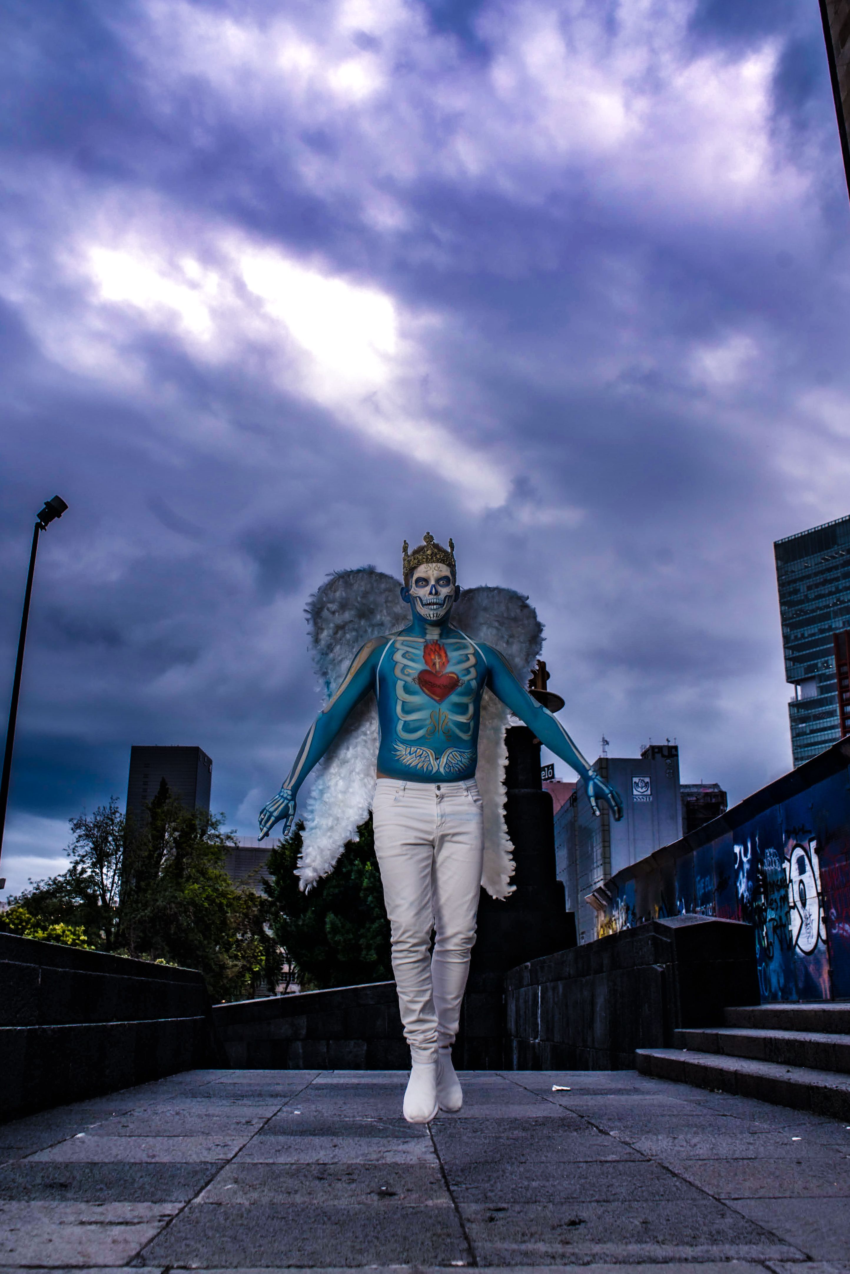 A striking street performer with skeleton body paint, angel wings, and a crown posing dramatically outdoors.