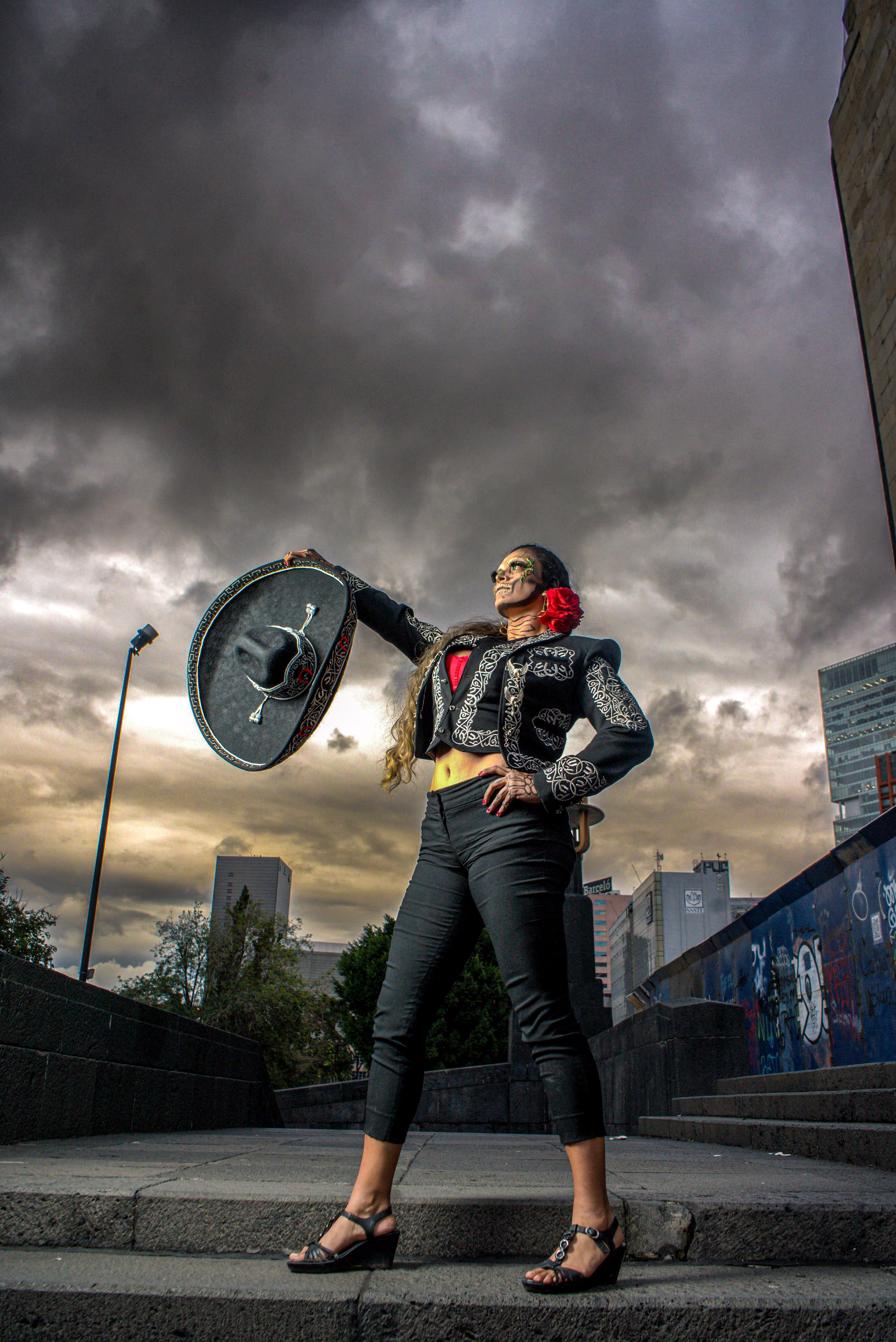 A woman poses boldly in traditional costume, holding a sombrero beneath a dramatic, cloudy sky in an urban setting.