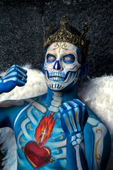 Close-up of a person with skull-themed body paint, wearing a crown and angel wings, set against an outdoor stone backdrop.
