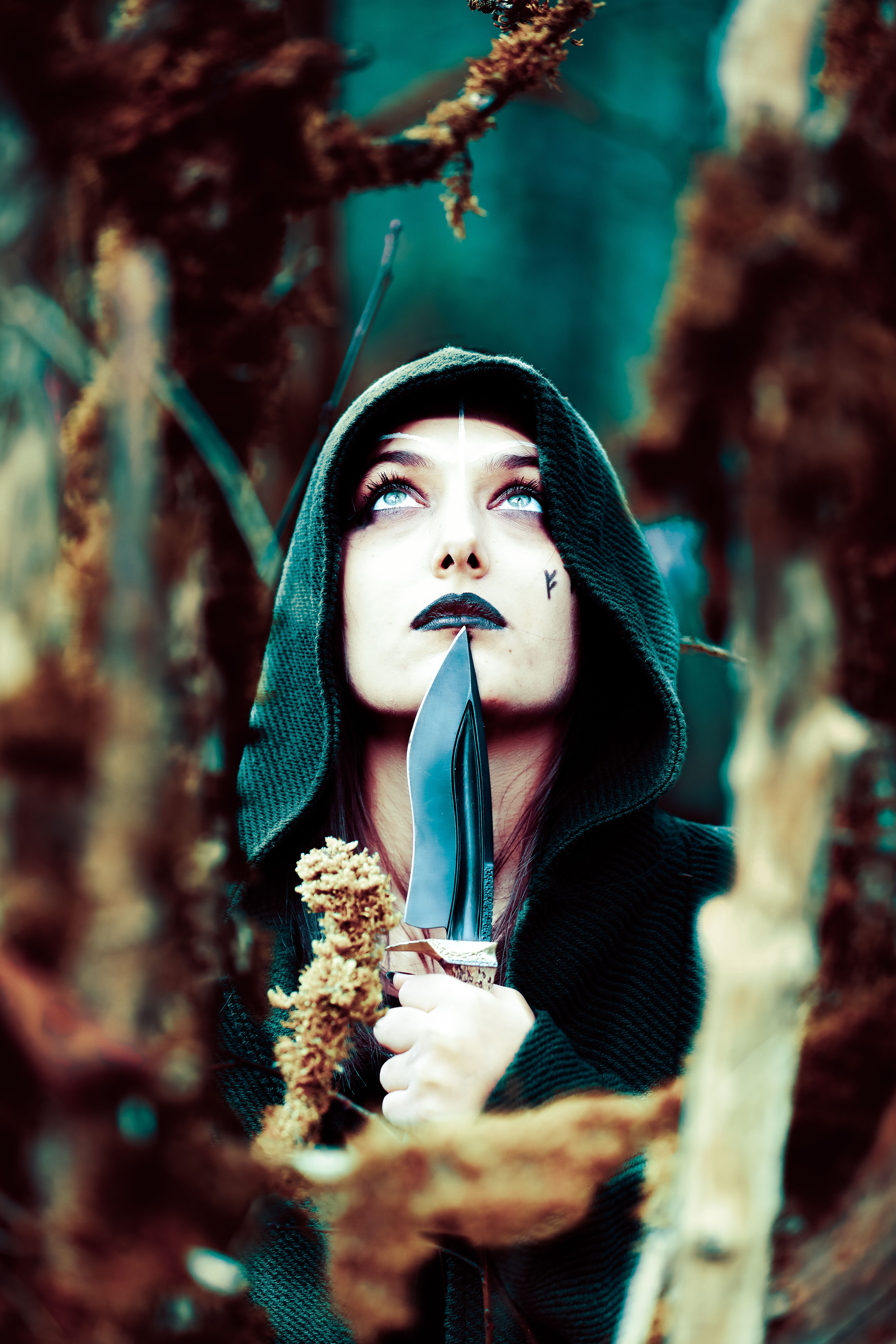 A person in a hooded cloak with dramatic makeup looks upwards holding a knife in a forest.