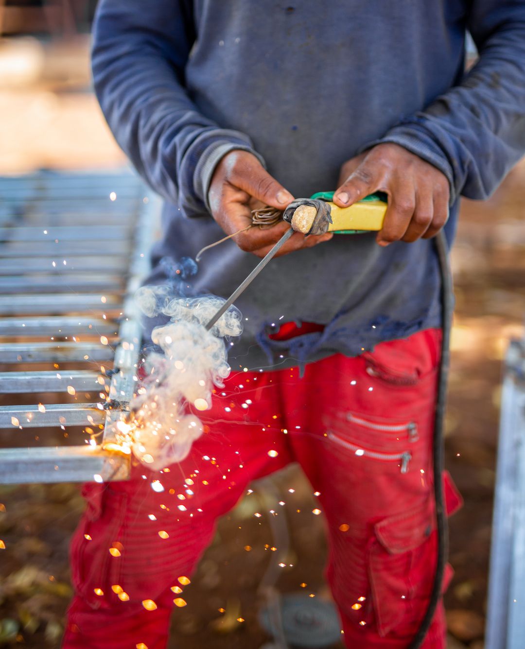 A man in work clothes cutting through a galvanized steel frame with a torch, orange sparks scattering across his bare hands and the metal
