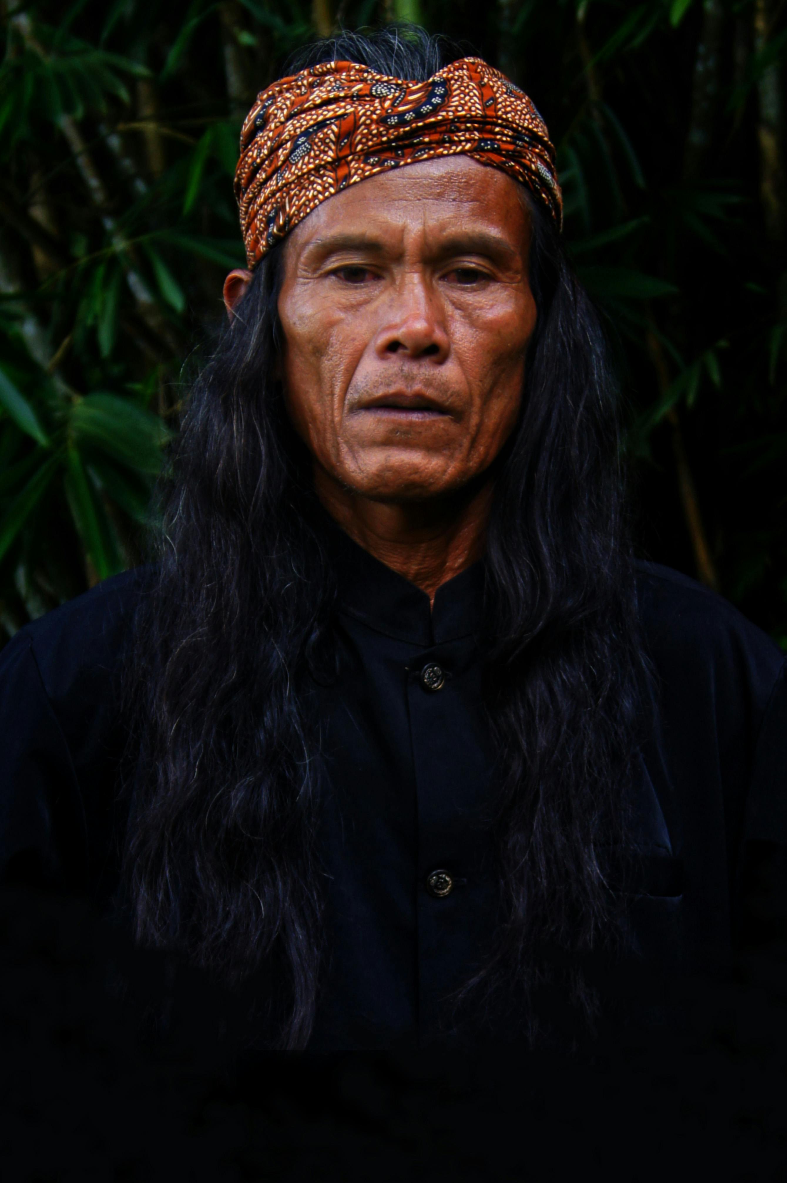 An adult man in traditional attire stands outdoors against a lush green backdrop, exuding mystery.