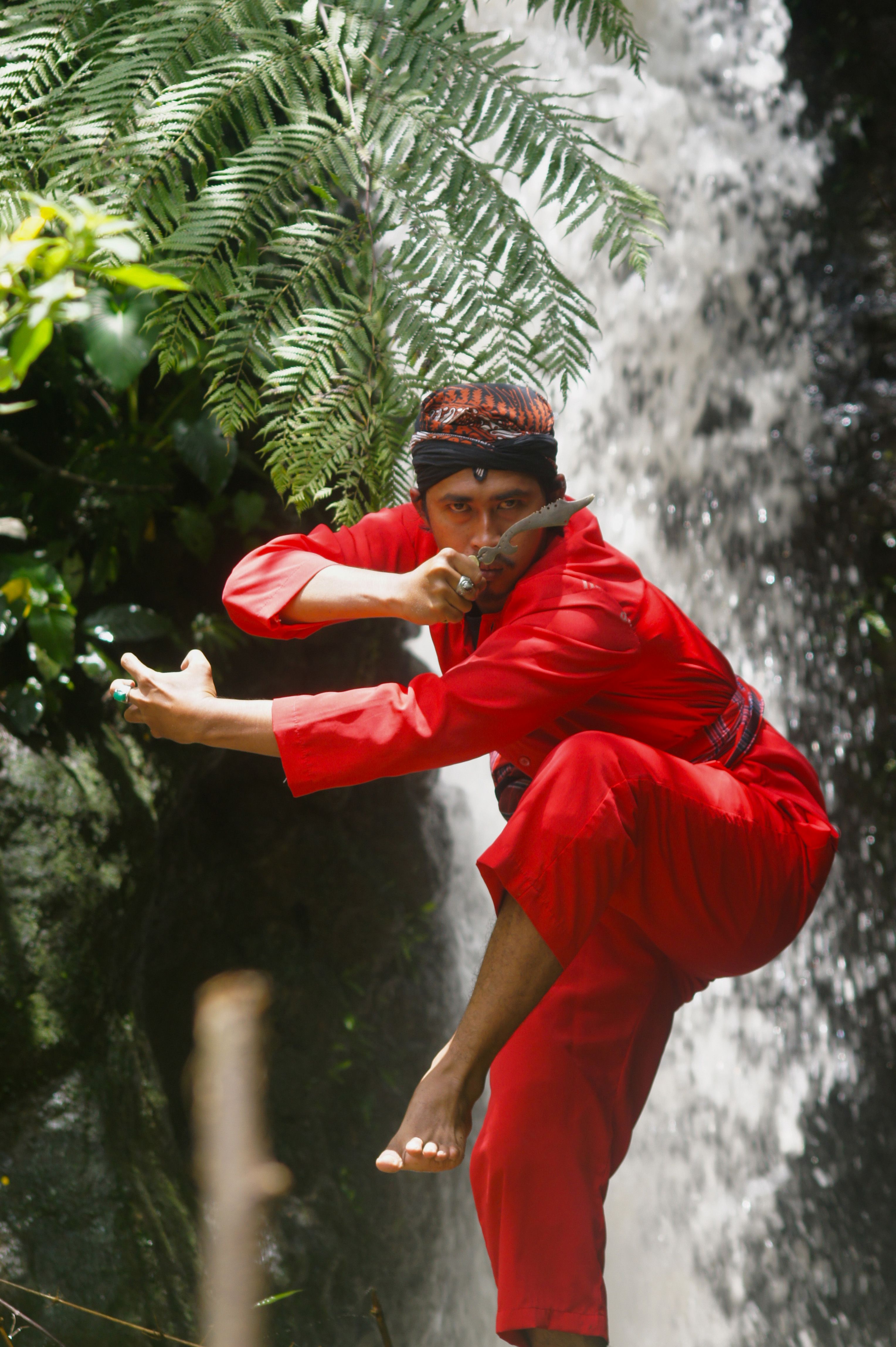 A martial artist in red attire performing a pose by a waterfall surrounded by ferns.