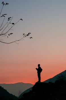 Silhouette of a person standing on a rock with mountains and sunset sky in the background, creating a tranquil scene.