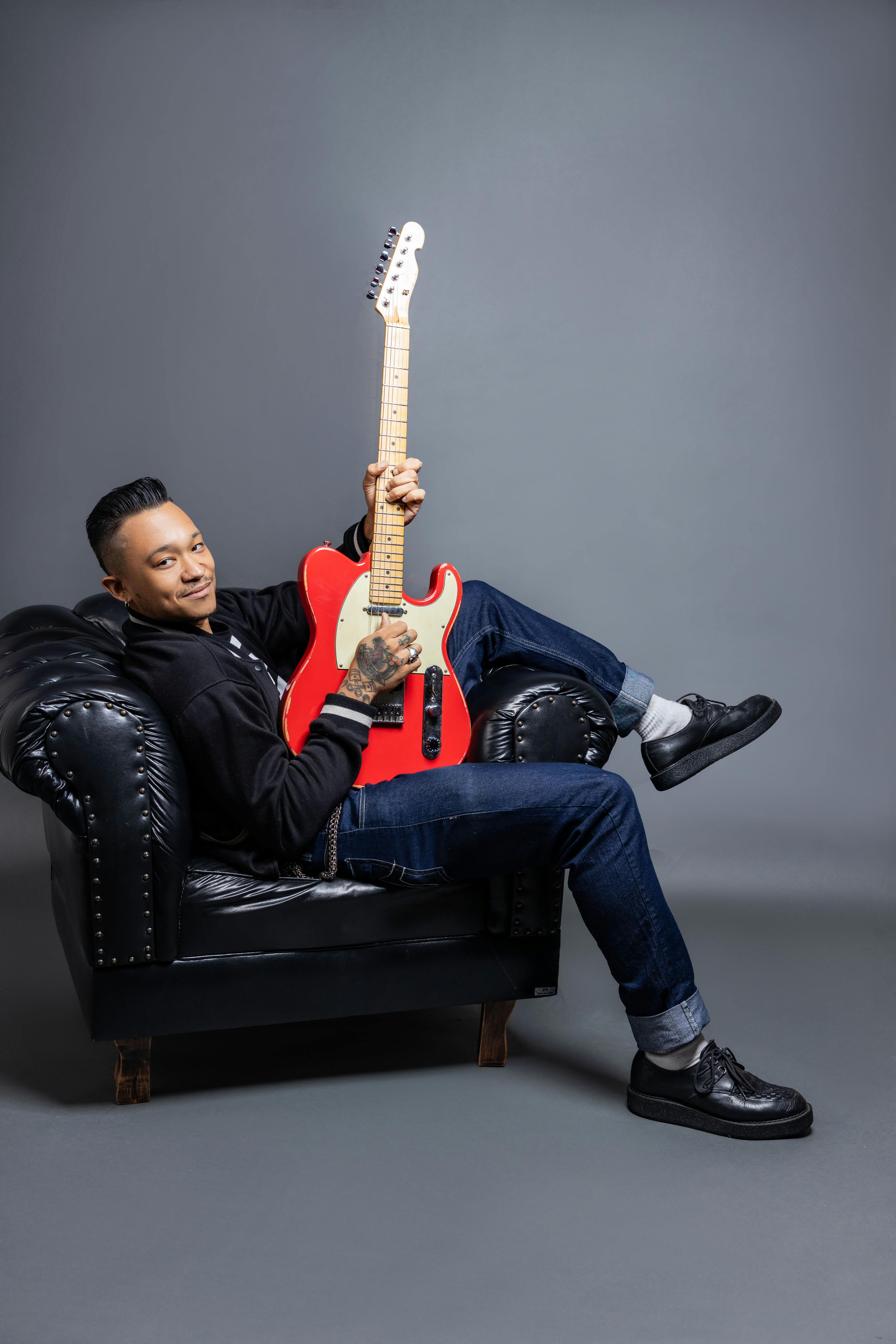 Man lounging on black sofa with a red electric guitar, in a studio setting.